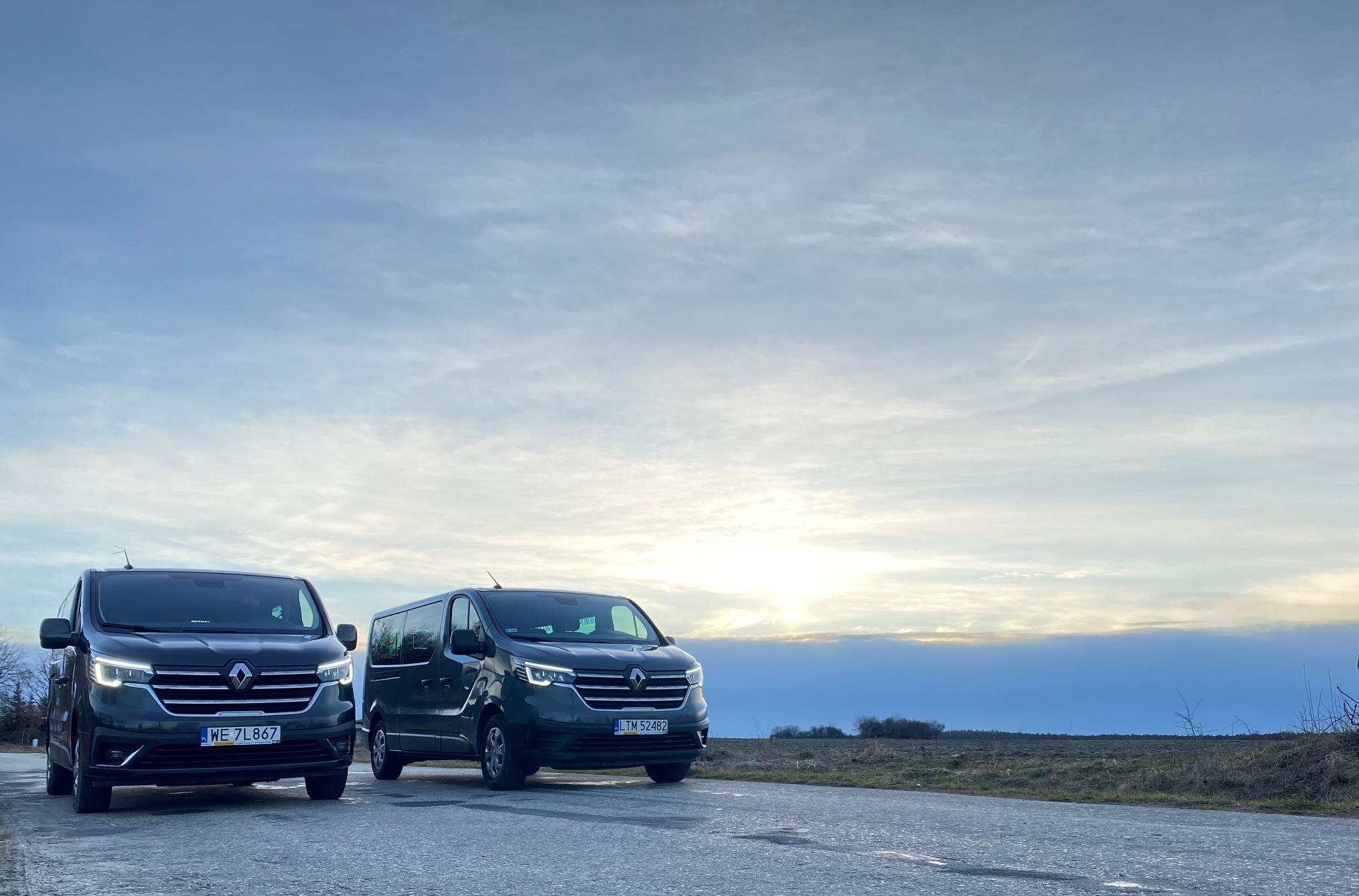 Dwa ciemnoszare busy Renault Trafic stojące na asfaltowej drodze na tle pochmurnego nieba o zachodzie słońca.