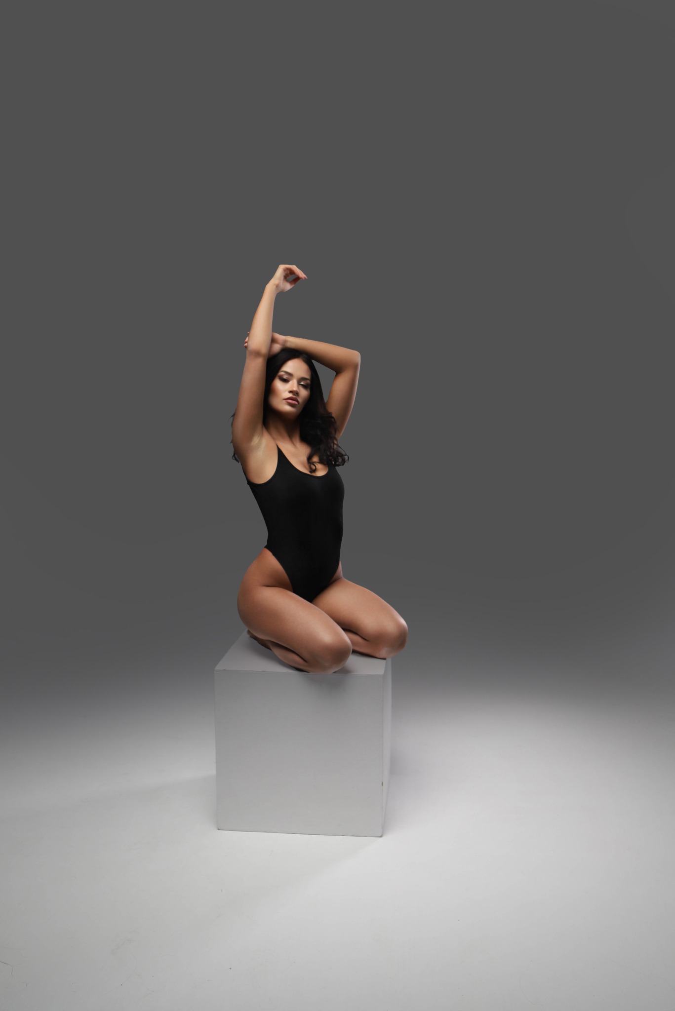 Kobieta w czarnym body pozuje, siedząc na białym sześcianie na szarym tle, z rękami uniesionymi nad głową.