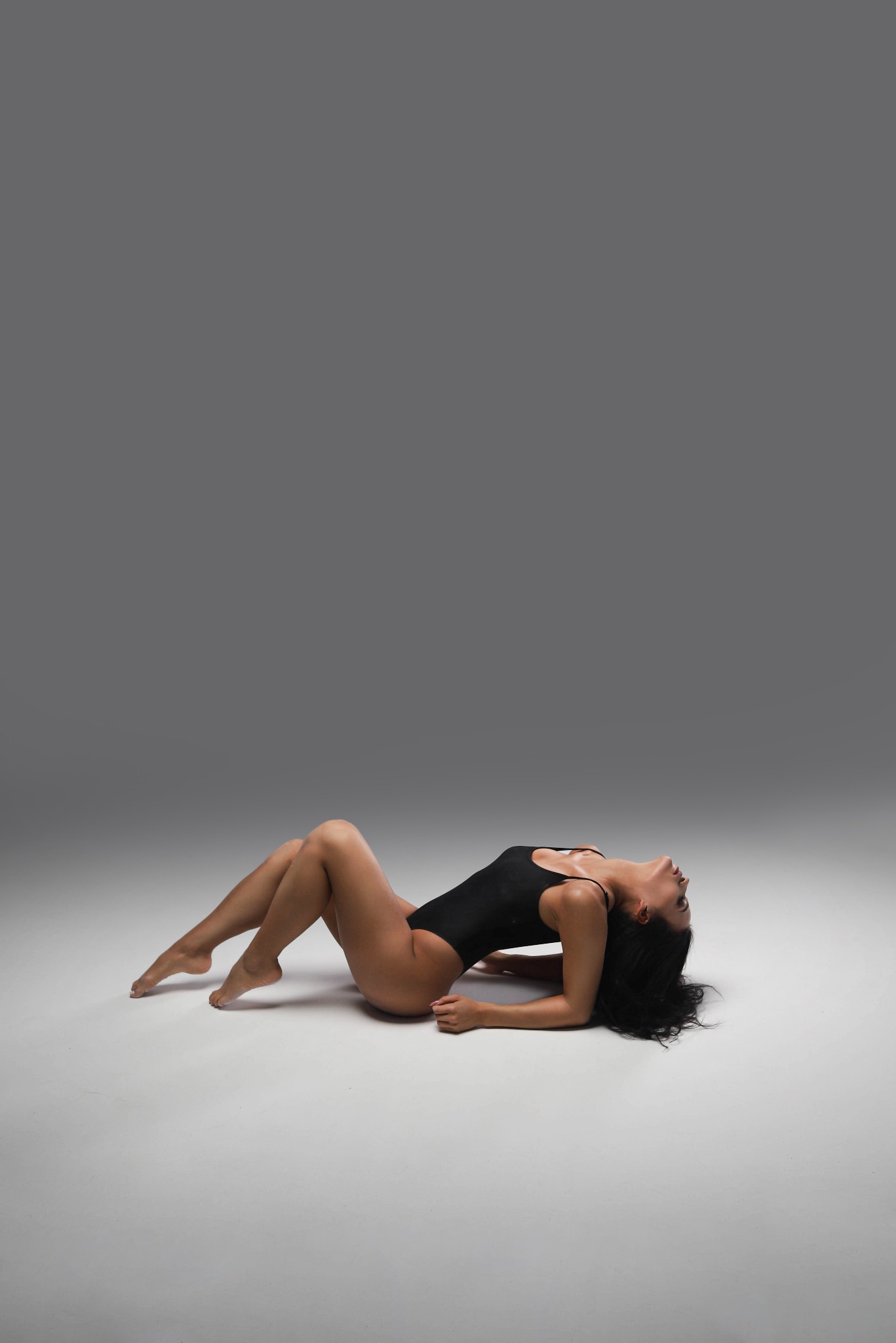 Kobieta w czarnym body leży na plecach na szarym tle, z głową odchyloną do tyłu i wygiętym ciałem.
