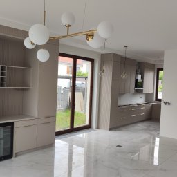 Usługi remontowo budowlane Kowalczyk Arkadiusz - Jasna kuchnia z beżowymi szafkami, marmurową podłogą i złotymi lampami wiszącymi. Widoczna zabudowa z barkiem i chłodziarką do wina.