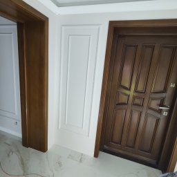 Usługi remontowo budowlane Kowalczyk Arkadiusz - Nowoczesne wnętrze po remoncie: brązowe drzwi wewnętrzne z panelową konstrukcją, białe ściany z dekoracyjnymi listwami, marmurowe płytki podłogowe z subtelnym żyłowaniem.