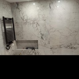 Usługi remontowo budowlane Kowalczyk Arkadiusz - Wykończona ściana łazienki z marmurową okładziną, wnęka z narzędziami i zainstalowany czarny grzejnik drabinkowy z elektrycznym podłączeniem.