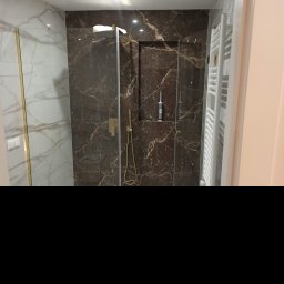 Usługi remontowo budowlane Kowalczyk Arkadiusz - Nowoczesna kabina prysznicowa z ciemnymi, marmurkowymi płytkami, złotą armaturą i szklaną ścianą, widoczny biały grzejnik drabinkowy i częściowo białe płytki na ścianie po lewej.