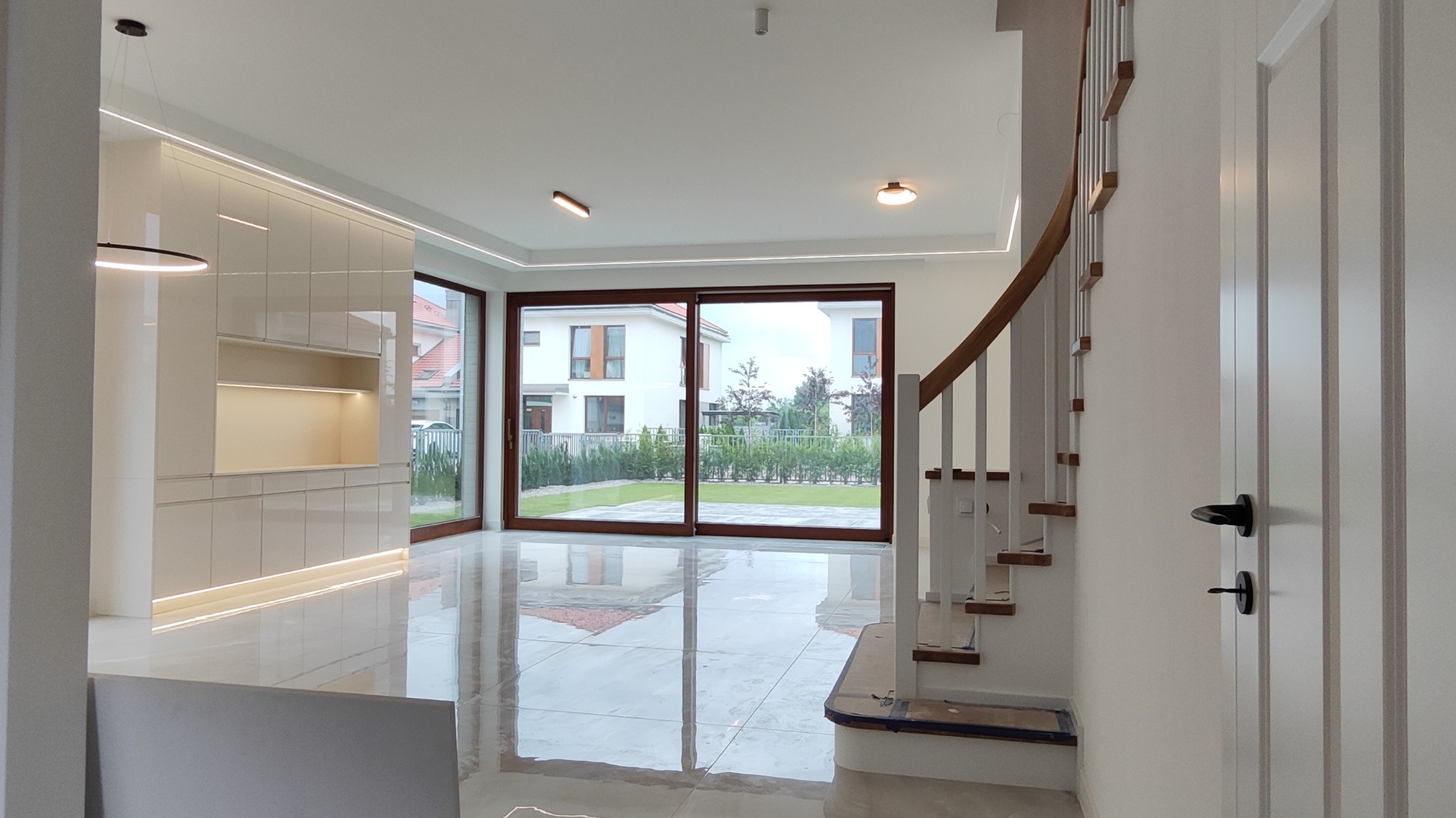 Jasne, przestronne wnętrze domu z widokiem na ogród przez duże przesuwne okna, z zabudową meblową z podświetleniem LED i schodami z drewnianymi stopniami.