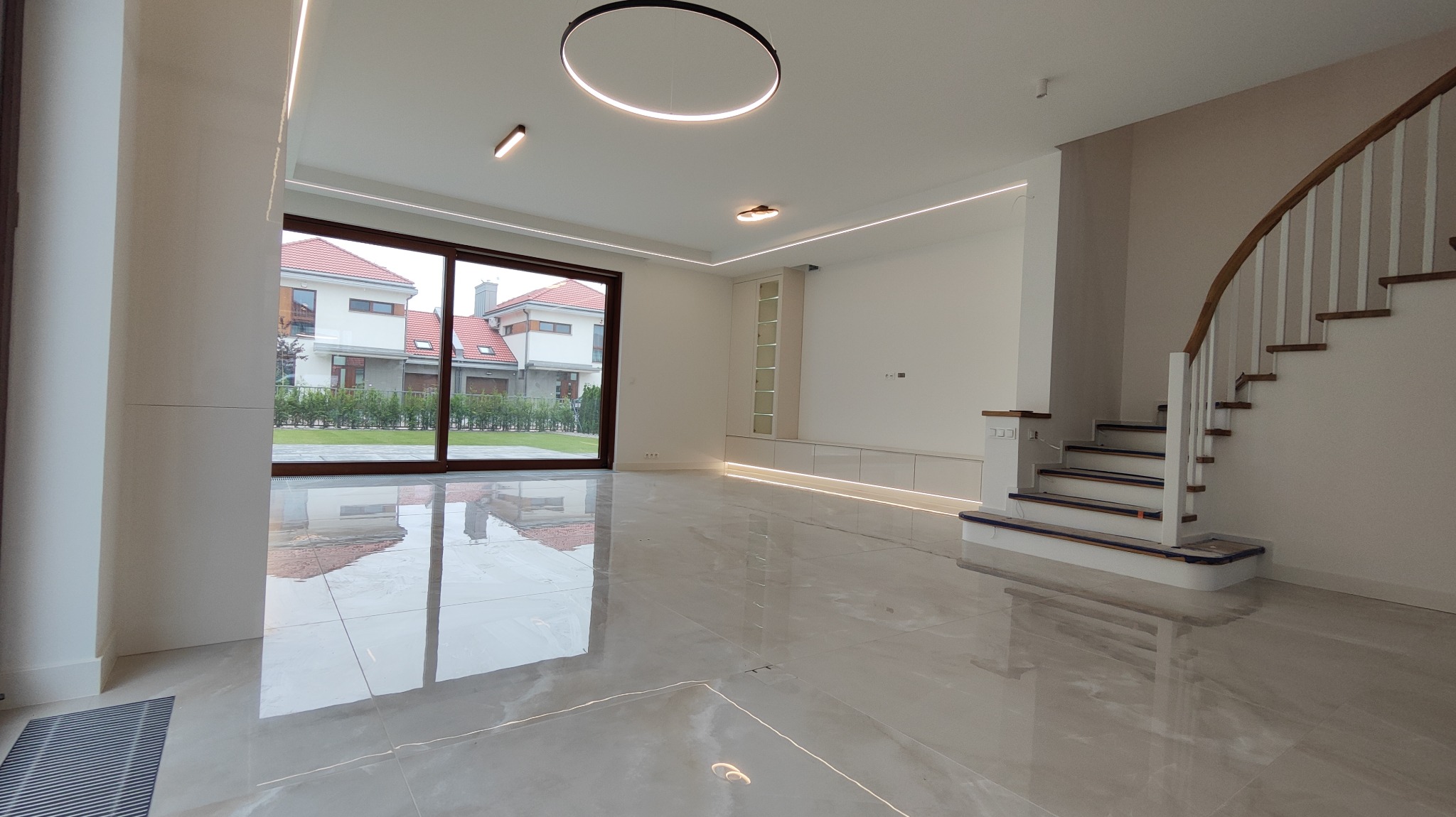 Przestronne, jasne wnętrze domu z marmurową podłogą, dużym oknem z widokiem na ogród, minimalistycznym oświetleniem sufitowym i drewnianymi schodami z białymi balustradami.