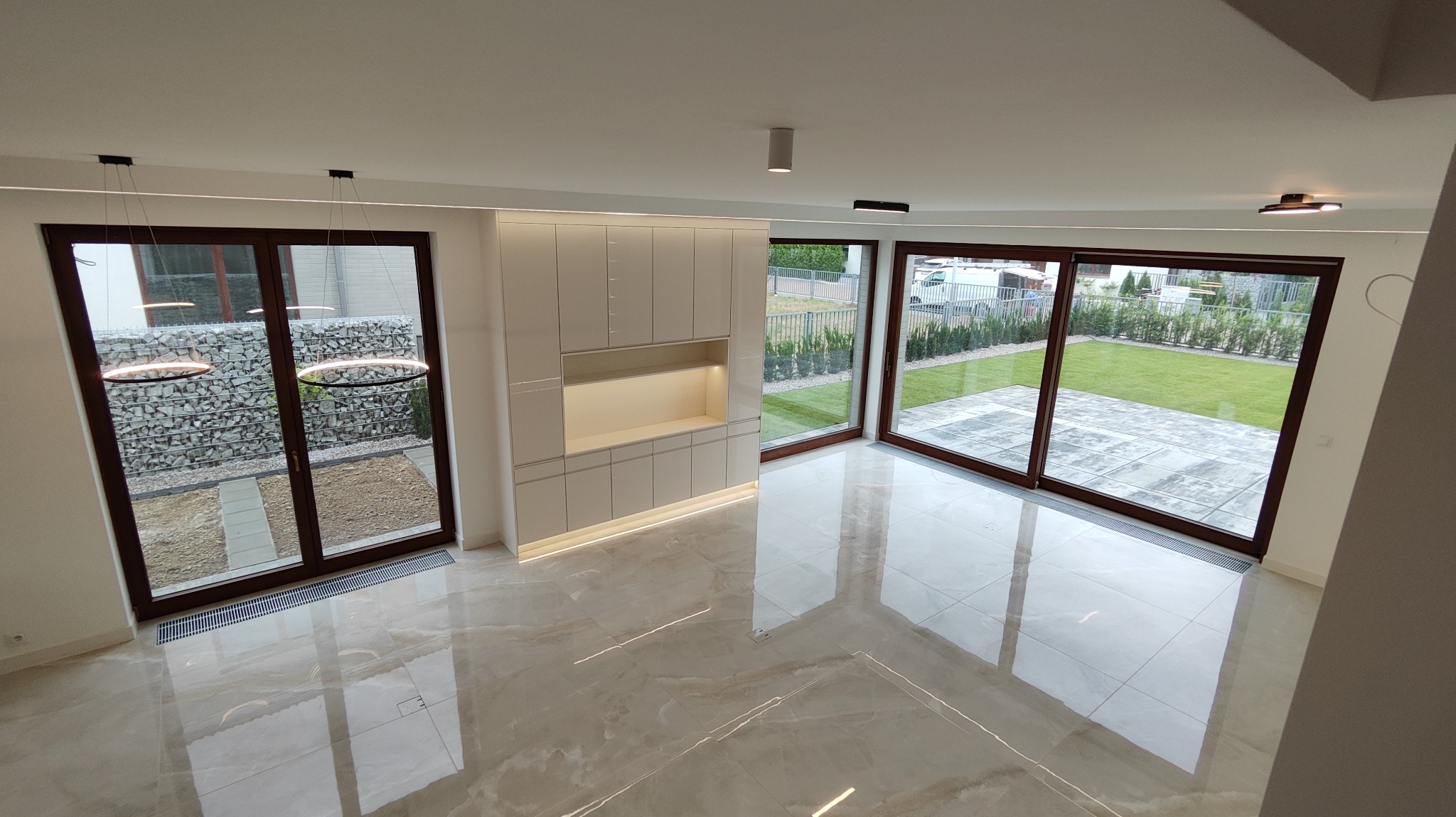 Nowoczesne wnętrze z błyszczącą marmurową podłogą, białymi szafkami z podświetleniem LED i dużymi przesuwnymi oknami z widokiem na ogród.