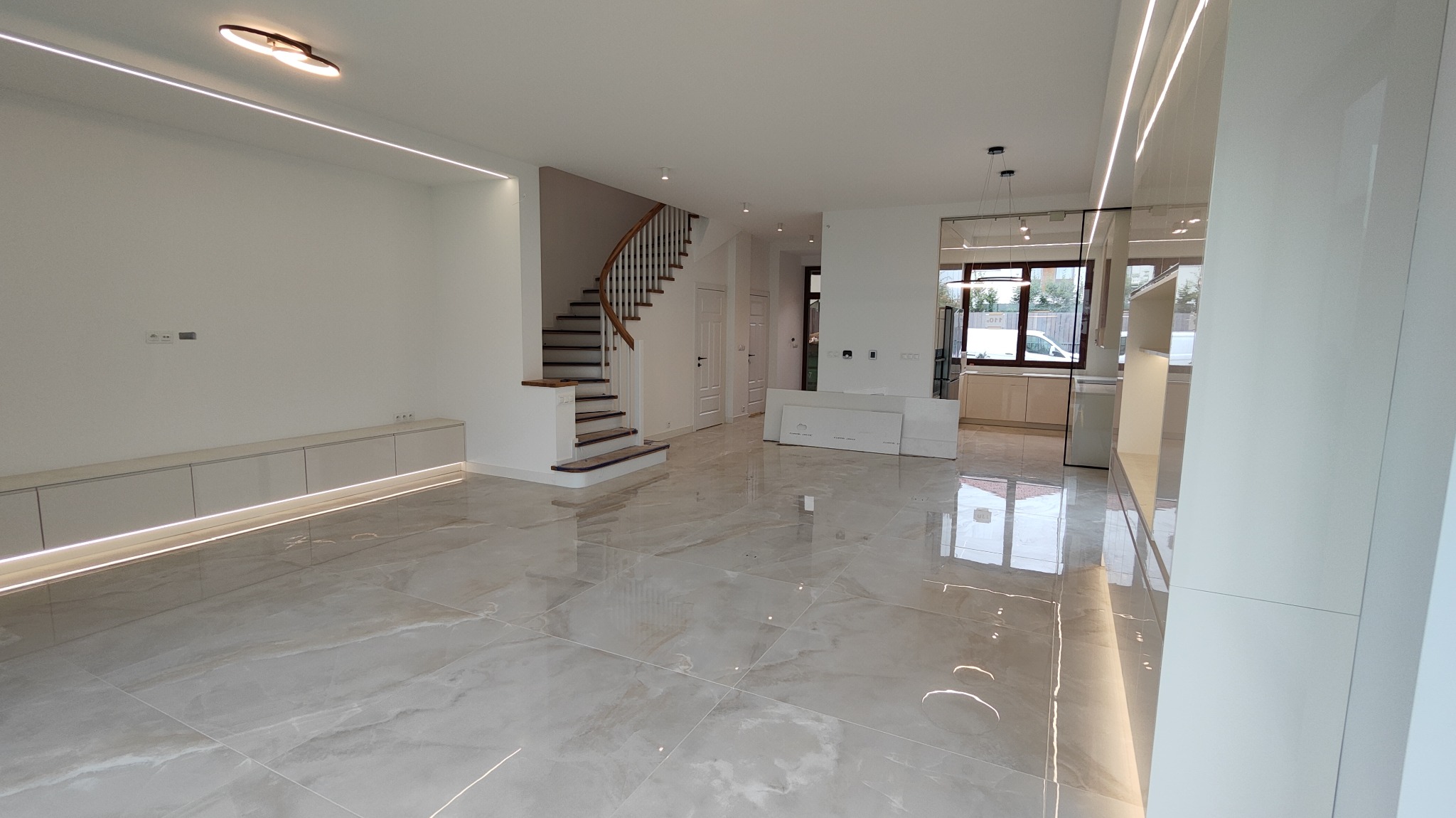Przestronne, jasne wnętrze domu po remoncie: marmurowe płytki podłogowe, schody z drewnianymi poręczami, białe ściany z oświetleniem LED i zabudowa meblowa w salonie z przejściem do kuchni.