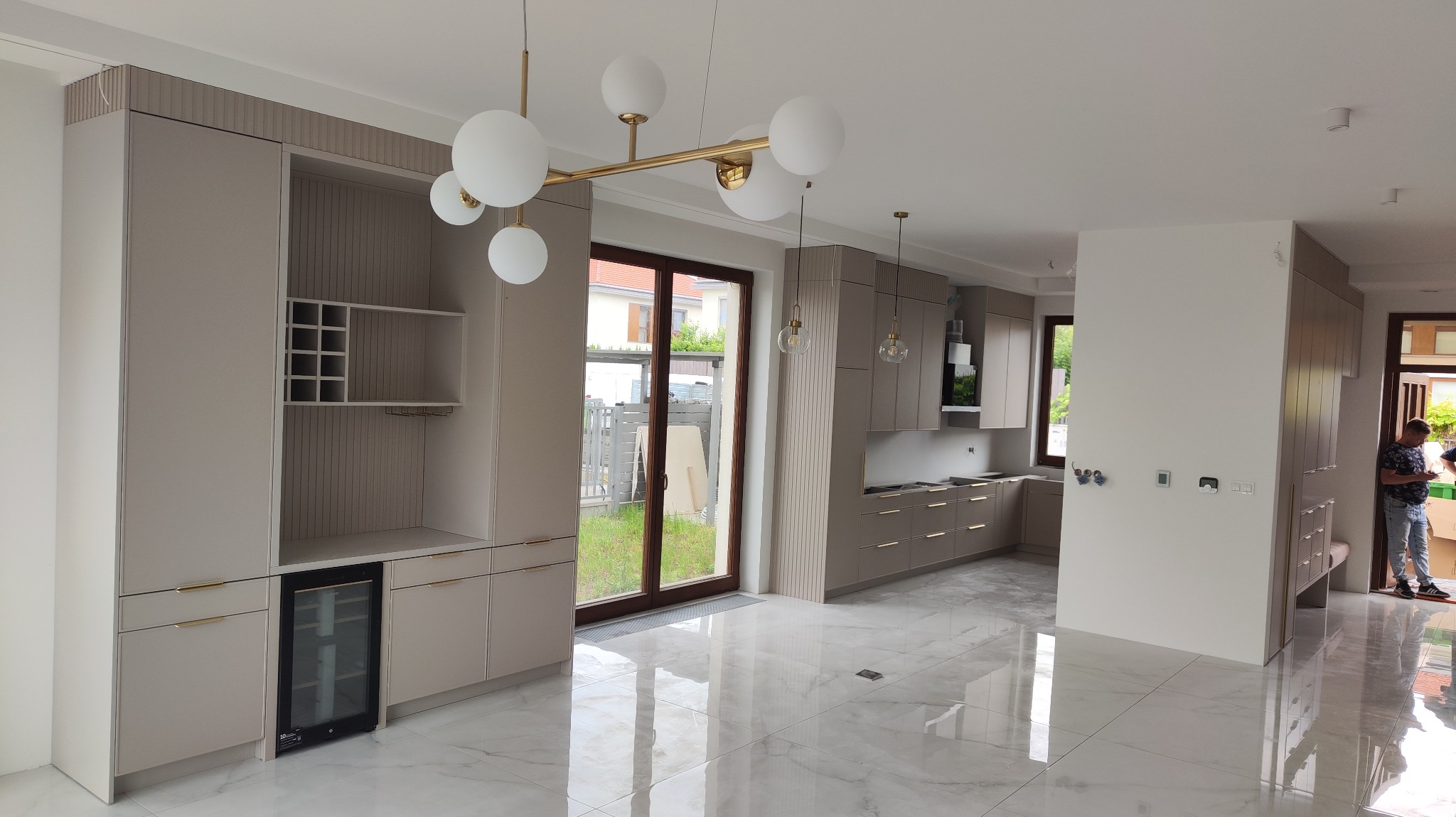 Jasna kuchnia z beżowymi szafkami, marmurową podłogą i złotymi lampami wiszącymi. Widoczna zabudowa z barkiem i chłodziarką do wina.