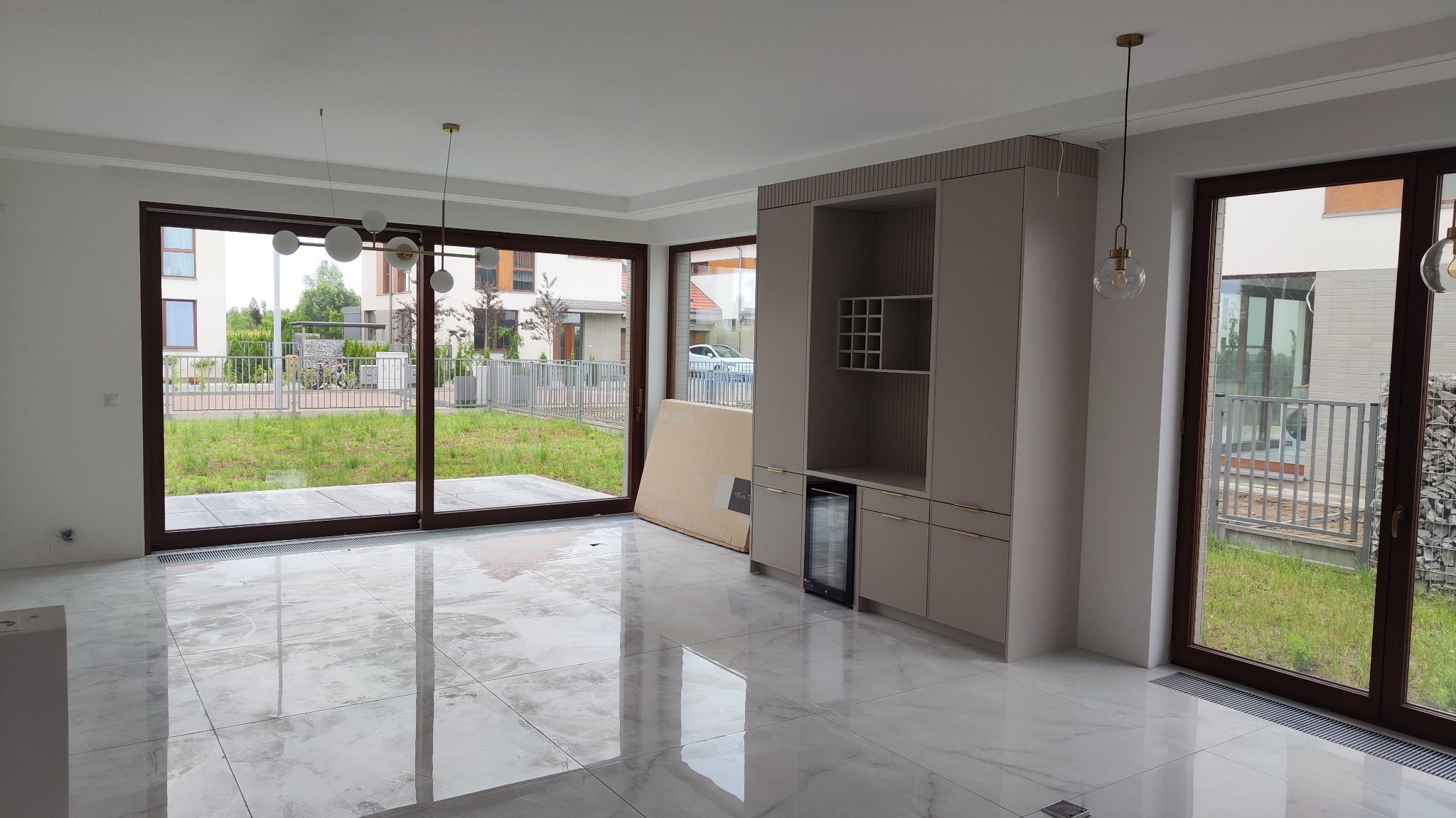 Wnętrze salonu w stanie deweloperskim z marmurową podłogą, dużymi oknami z widokiem na ogród, zabudową meblową z barkiem i wiszącymi lampami kulami.