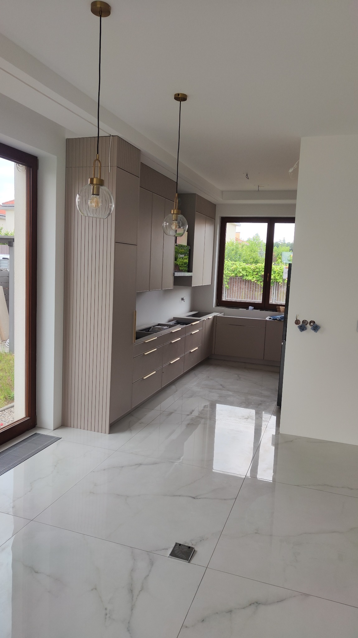 Jasna kuchnia z marmurową podłogą i szarymi szafkami z poziomymi żłobieniami, złote uchwyty i lampy wiszące z przezroczystymi kloszami, widok na zieleń przez okno.
