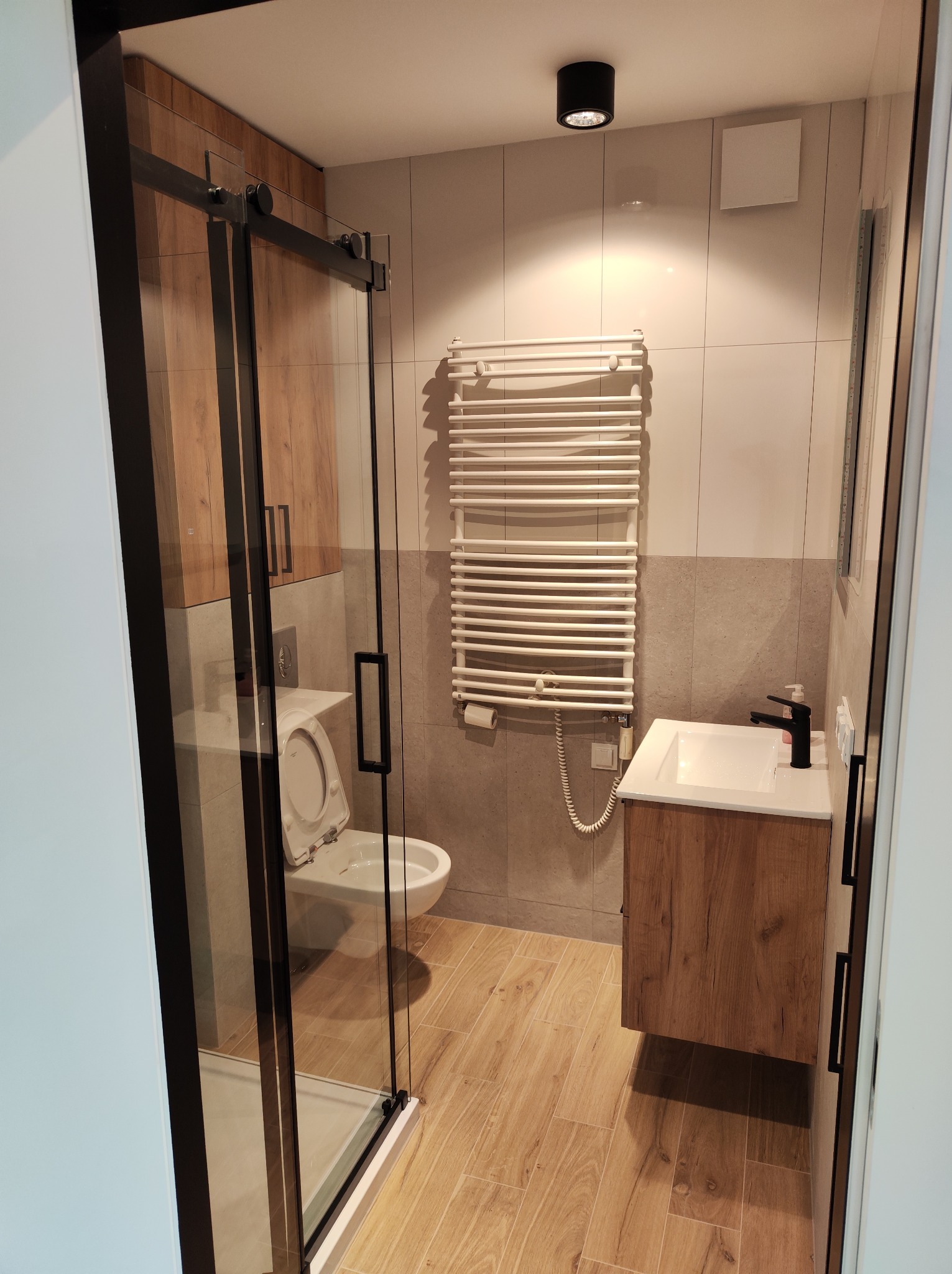 Nowoczesna łazienka z kabiną prysznicową z czarnymi ramami, drewnopodobnymi szafkami i grzejnikiem drabinkowym na tle szaro-białych płytek.