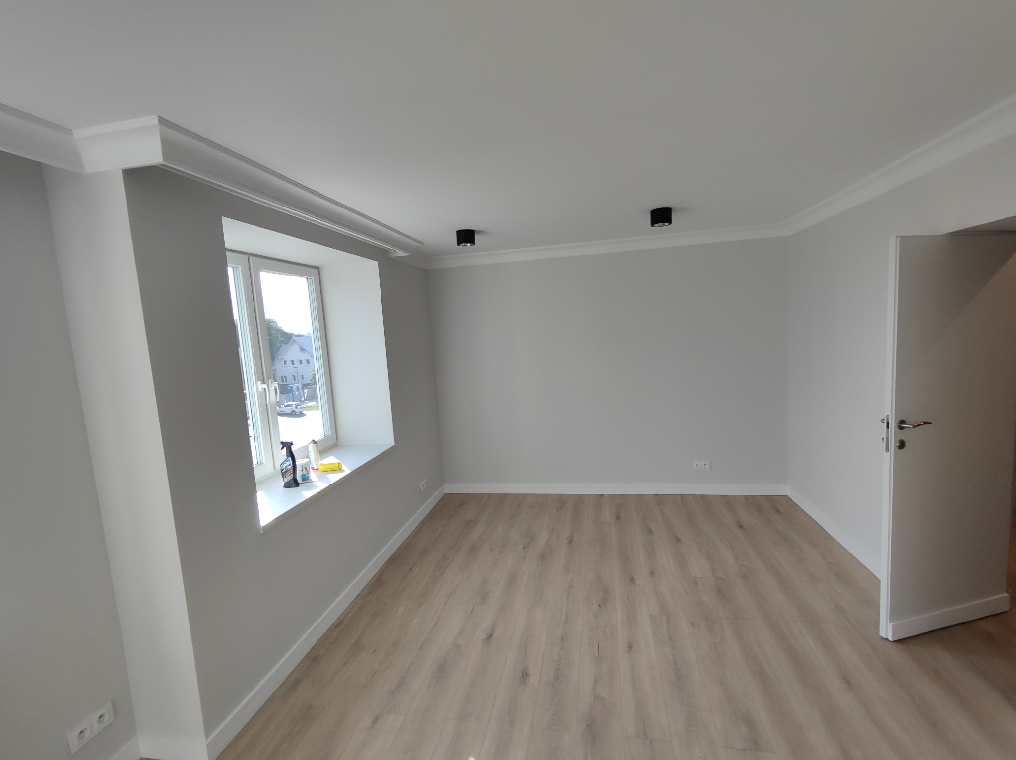 Jasne, puste pomieszczenie z panelami podłogowymi, listwami przypodłogowymi, ozdobnymi listwami sufitowymi, oknem z widokiem na zewnątrz i otwartymi drzwiami, gotowe do umeblowania.