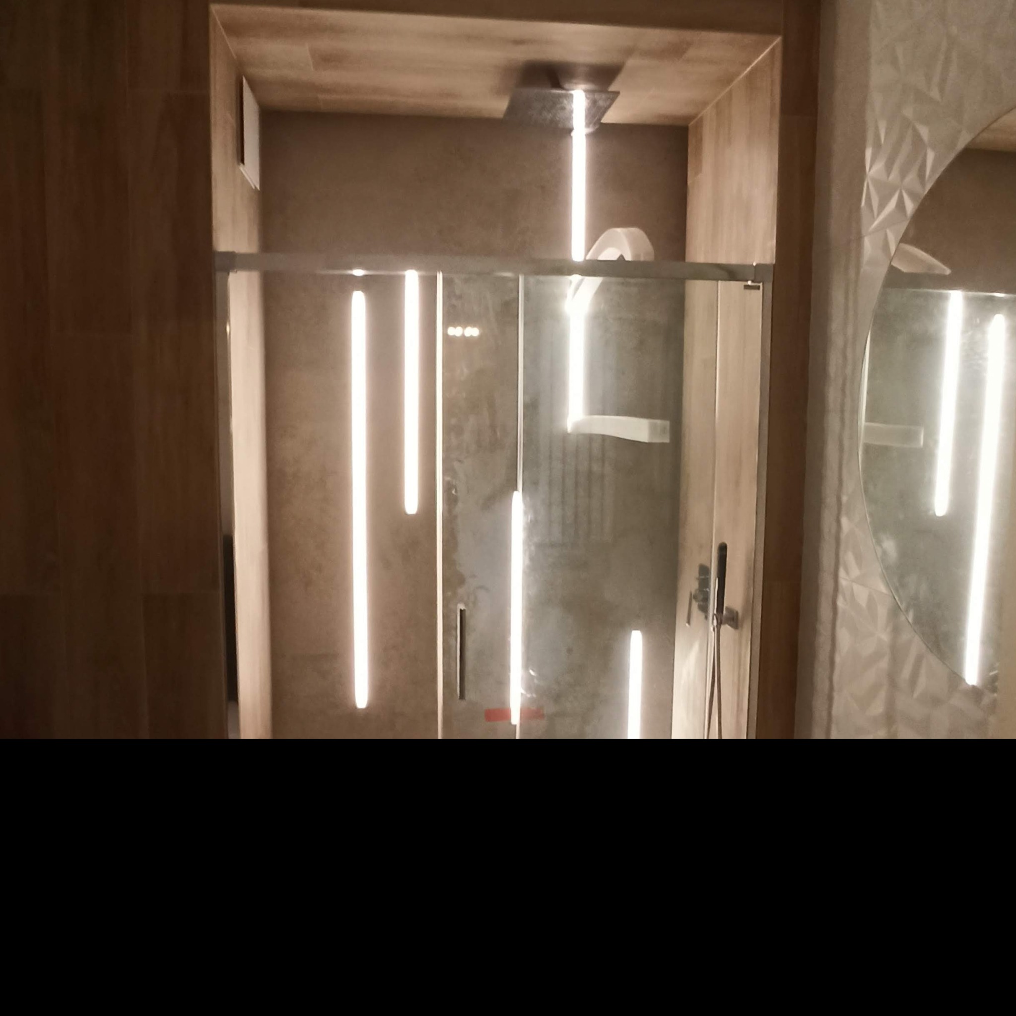 Nowoczesna kabina prysznicowa z oświetleniem LED w minimalistycznej łazience z dekoracyjną ścianą i okrągłym lustrem.