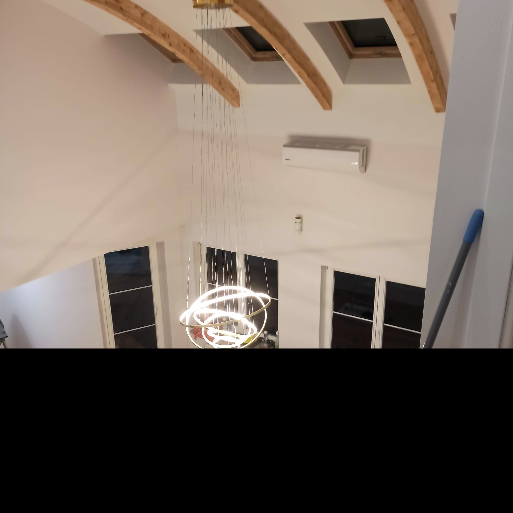 Wysoki pokój na poddaszu z drewnianymi belkami stropowymi, oknami dachowymi, klimatyzacją i nowoczesnym, okrągłym żyrandolem z cienkimi przewodami.