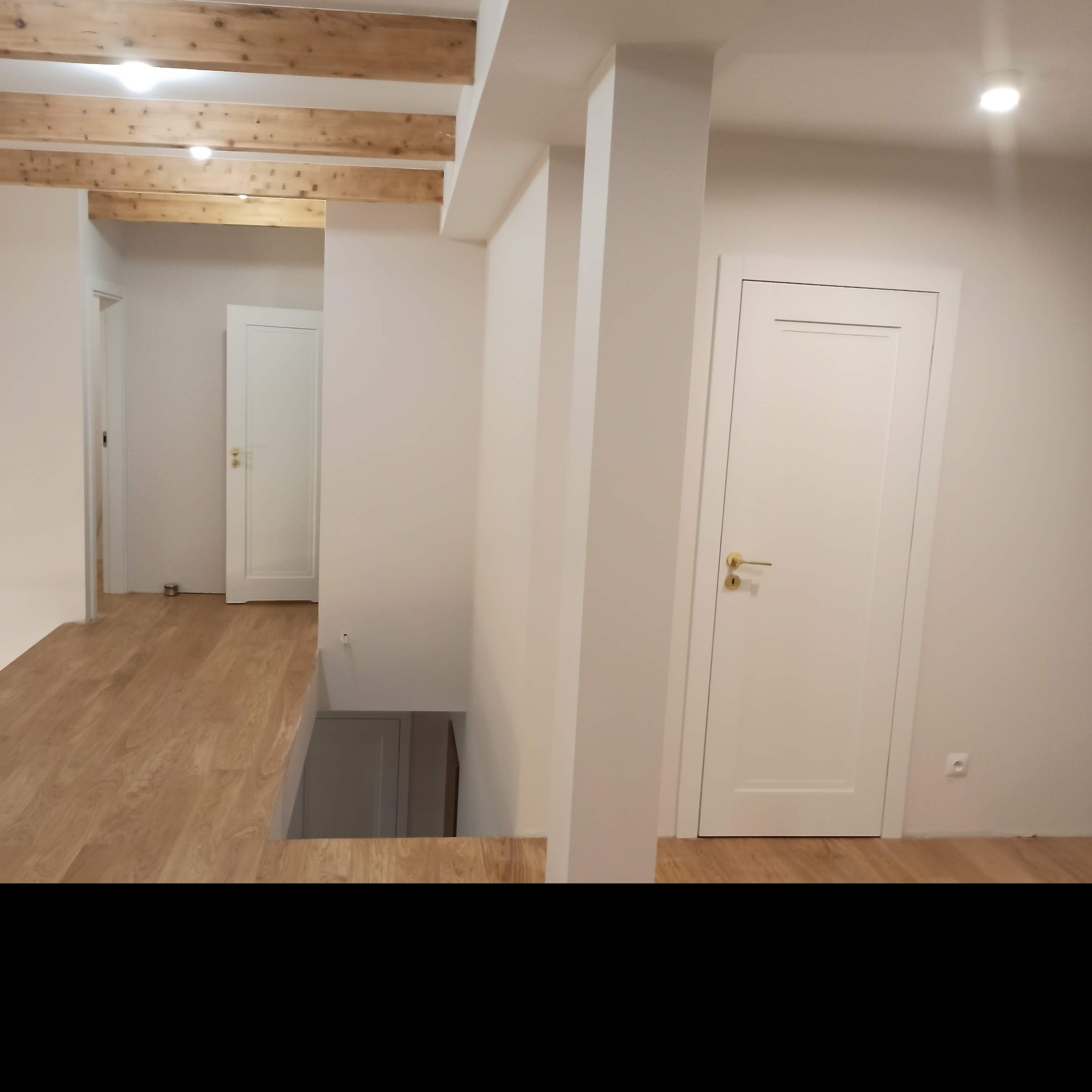 Jasne wnętrze z drewnianą podłogą, białymi ścianami, kolumną i drewnianymi belkami stropowymi, widoczne białe drzwi z złotymi klamkami.