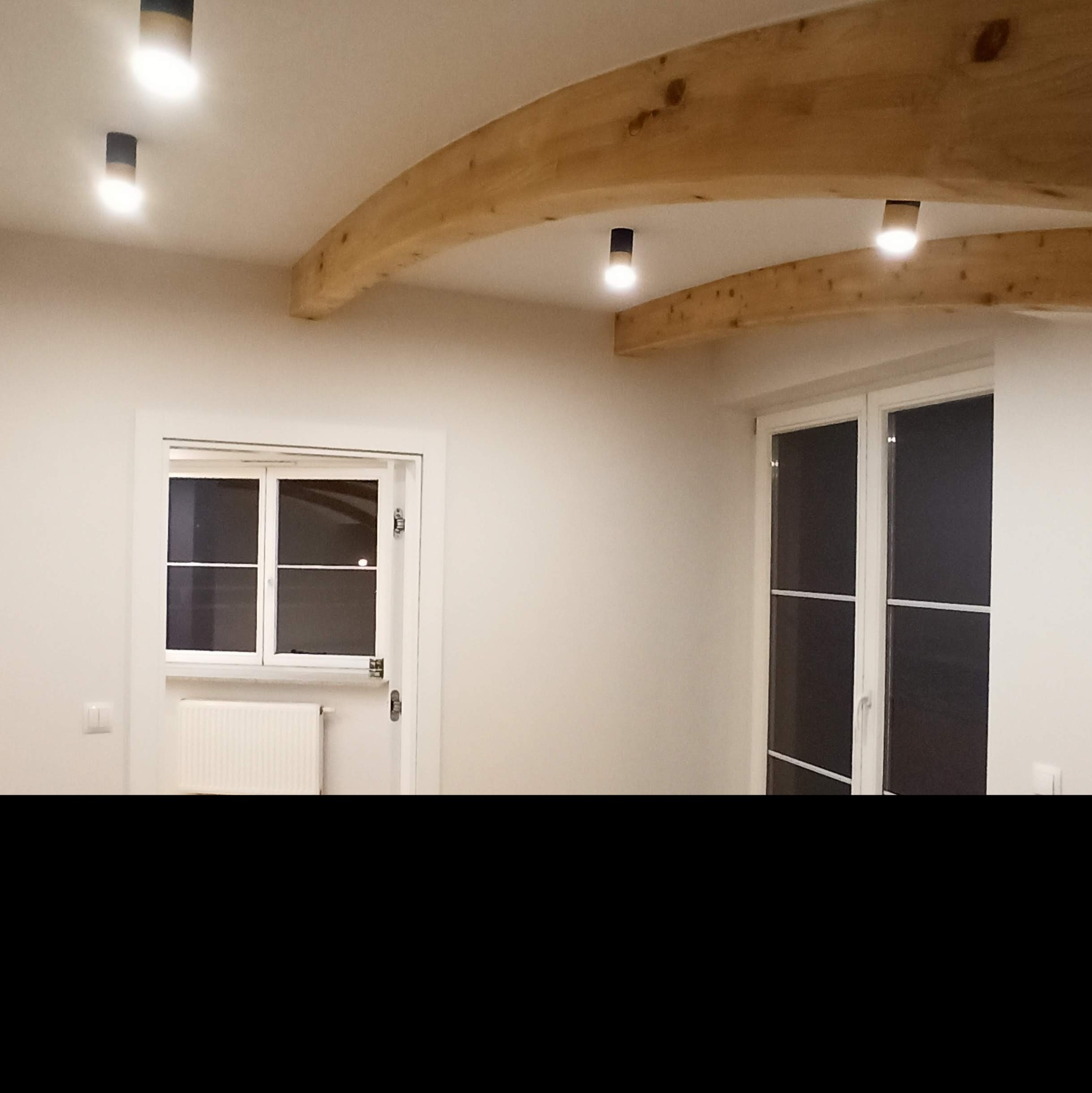 Jasne wnętrze z drewnianymi belkami stropowymi, punktowym oświetleniem sufitowym, białymi ścianami, oknem z grzejnikiem pod spodem i drugim oknem z moskitierą.