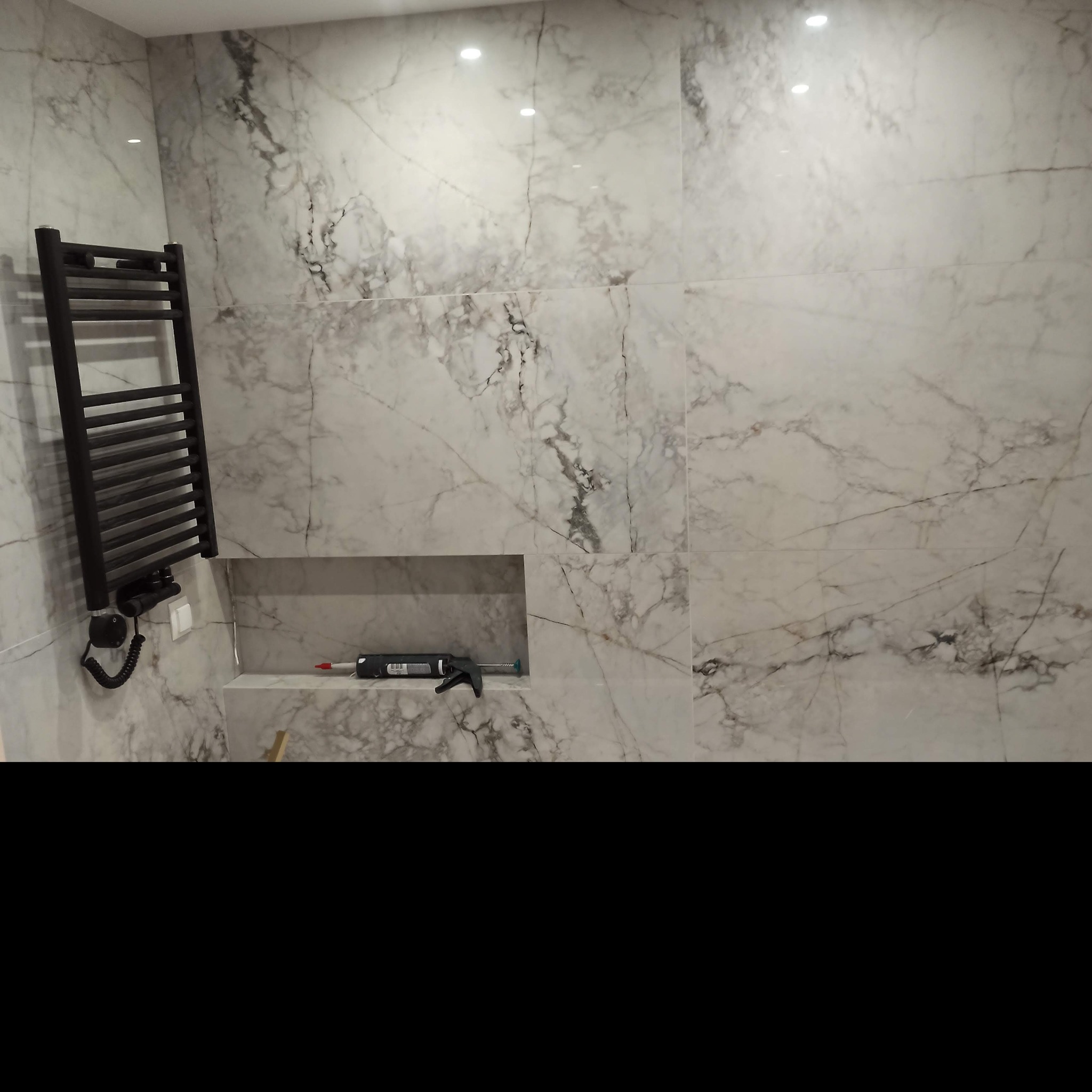 Wykończona ściana łazienki z marmurową okładziną, wnęka z narzędziami i zainstalowany czarny grzejnik drabinkowy z elektrycznym podłączeniem.