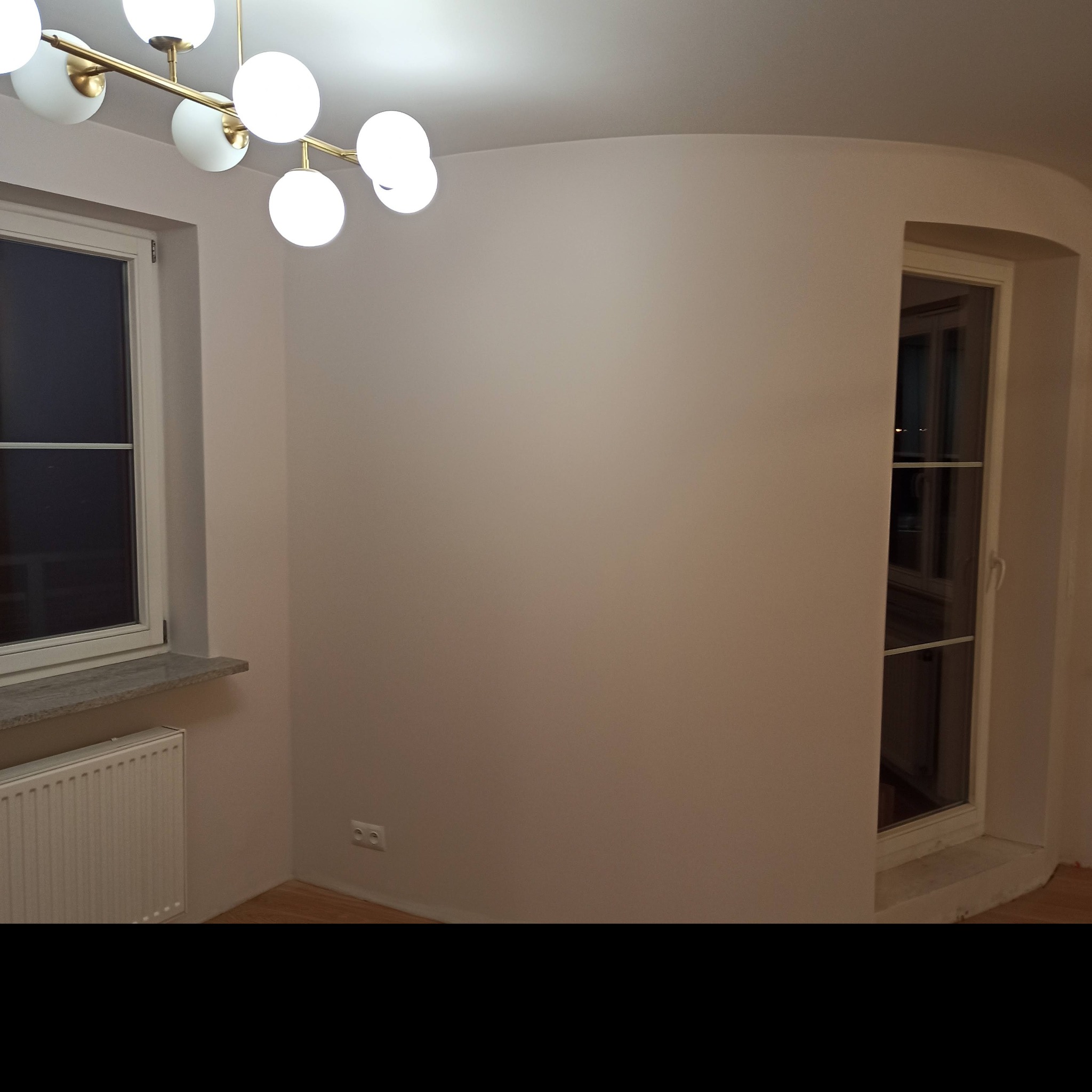 Pomalowane na jasny kolor wnętrze z oknami, grzejnikiem i nowoczesną lampą sufitową z kulistymi kloszami. Ściana po prawej stronie z zaokrąglonym narożnikiem przy oknie.