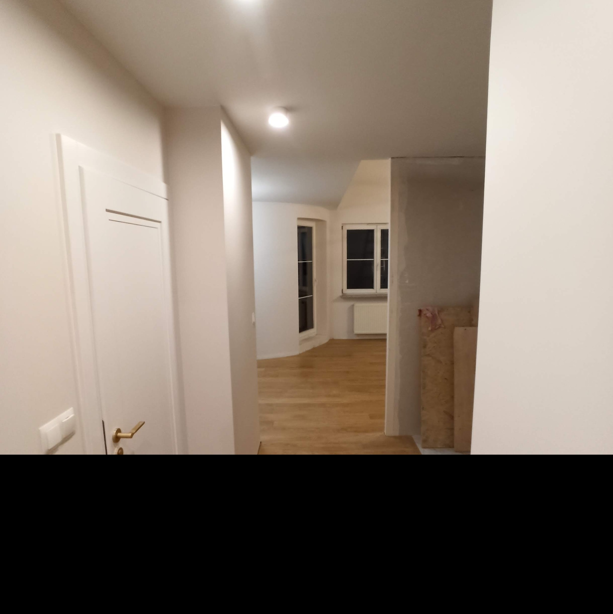 Korytarz w trakcie wykańczania, widoczne białe drzwi z złotą klamką, świeżo położona drewniana podłoga, ściany pomalowane na biało, w głębi pokój z oknami i grzejnikiem, fragment ściany...