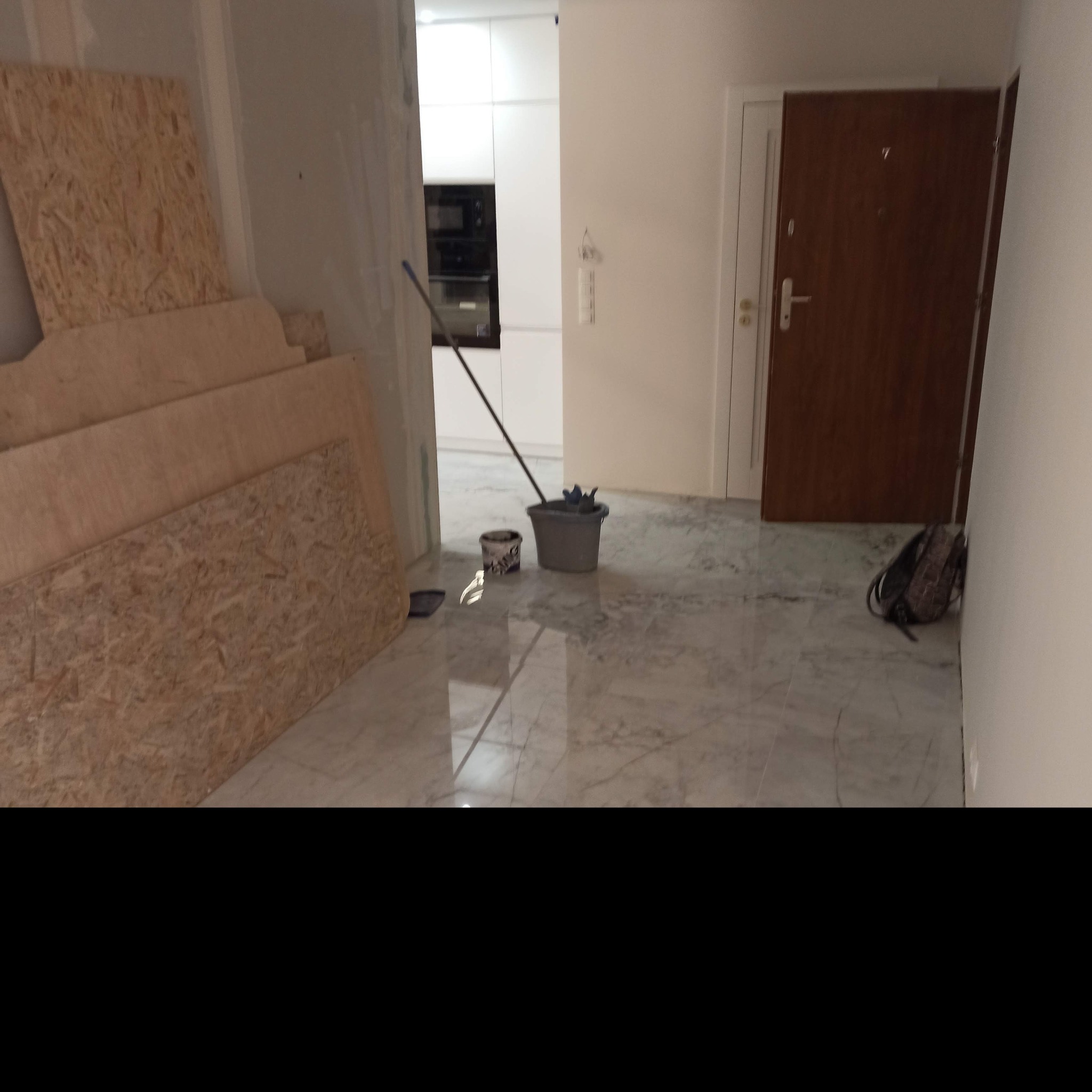 Przygotowania do remontu mieszkania: płyty OSB o różnych rozmiarach, wiadro z mopem, puszka po farbie i plecak w przedpokoju z marmurową podłogą, widok na kuchnię.