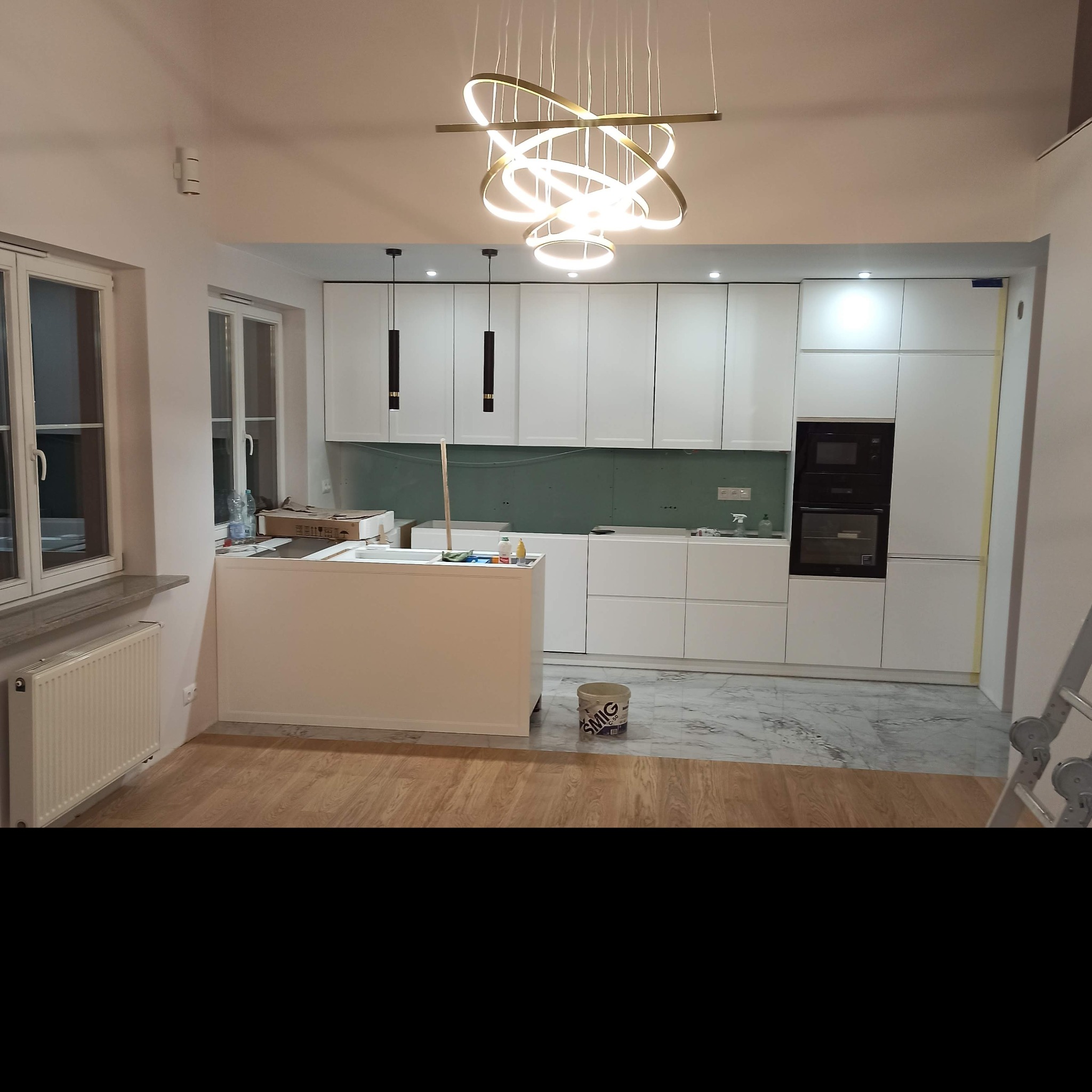 Nowoczesna, biała kuchnia w trakcie montażu, z wyspą i designerską lampą wiszącą, widoczny karton gips na ścianie i wiadro z klejem na podłodze.