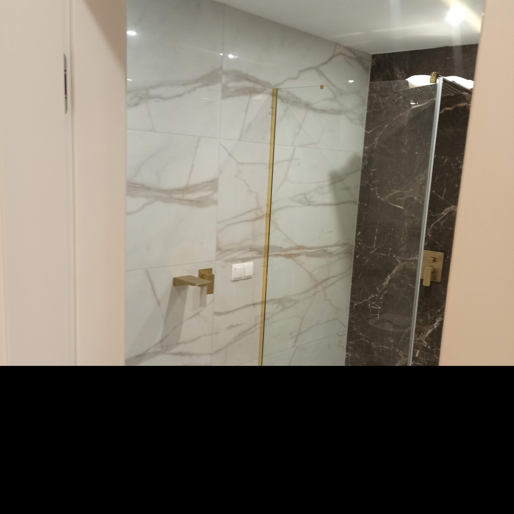 Nowoczesna łazienka z marmurowymi płytkami w odcieniach bieli i brązu, złota armatura i kabina prysznicowa z ciemnego marmuru.