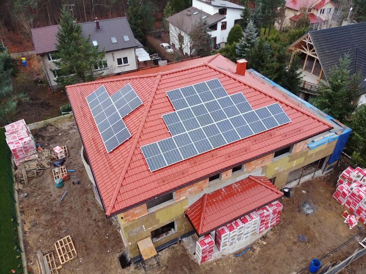 Dom w trakcie budowy z zamontowanymi panelami słonecznymi na czerwonym dachu, widok z góry, materiały budowlane składowane obok.