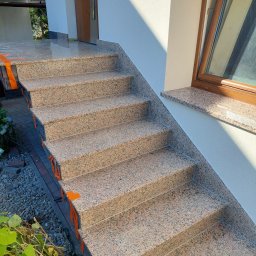 Paweł Mierzwa - Zewnętrzne schody wejściowe wyłożone płytami z różowego granitu, z widocznymi pomarańczowymi elementami zabezpieczającymi krawędzie, prowadzące do drzwi wejściowych i okna z drewnianą ramą.