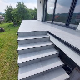 Paweł Mierzwa - Zewnętrzne schody i taras wyłożone jasnoszarą płytką ceramiczną z czarnymi fugami, widok z dołu na tle zielonego trawnika i drzew