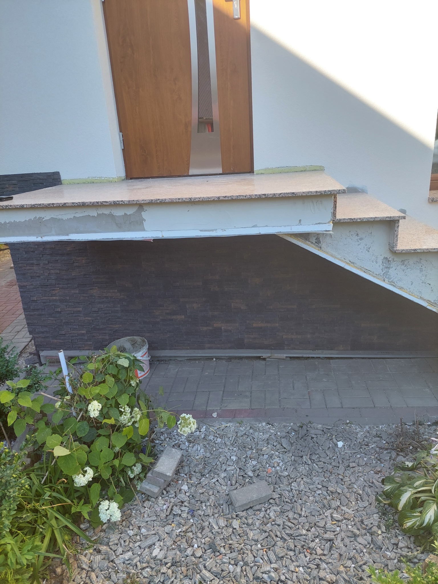 Montaż okładziny z cegły klinkierowej na podmurówce schodów wejściowych, widoczny częściowo ukończony etap prac z pozostawionym wiadrem po zaprawie i elementami roślinności w tle.