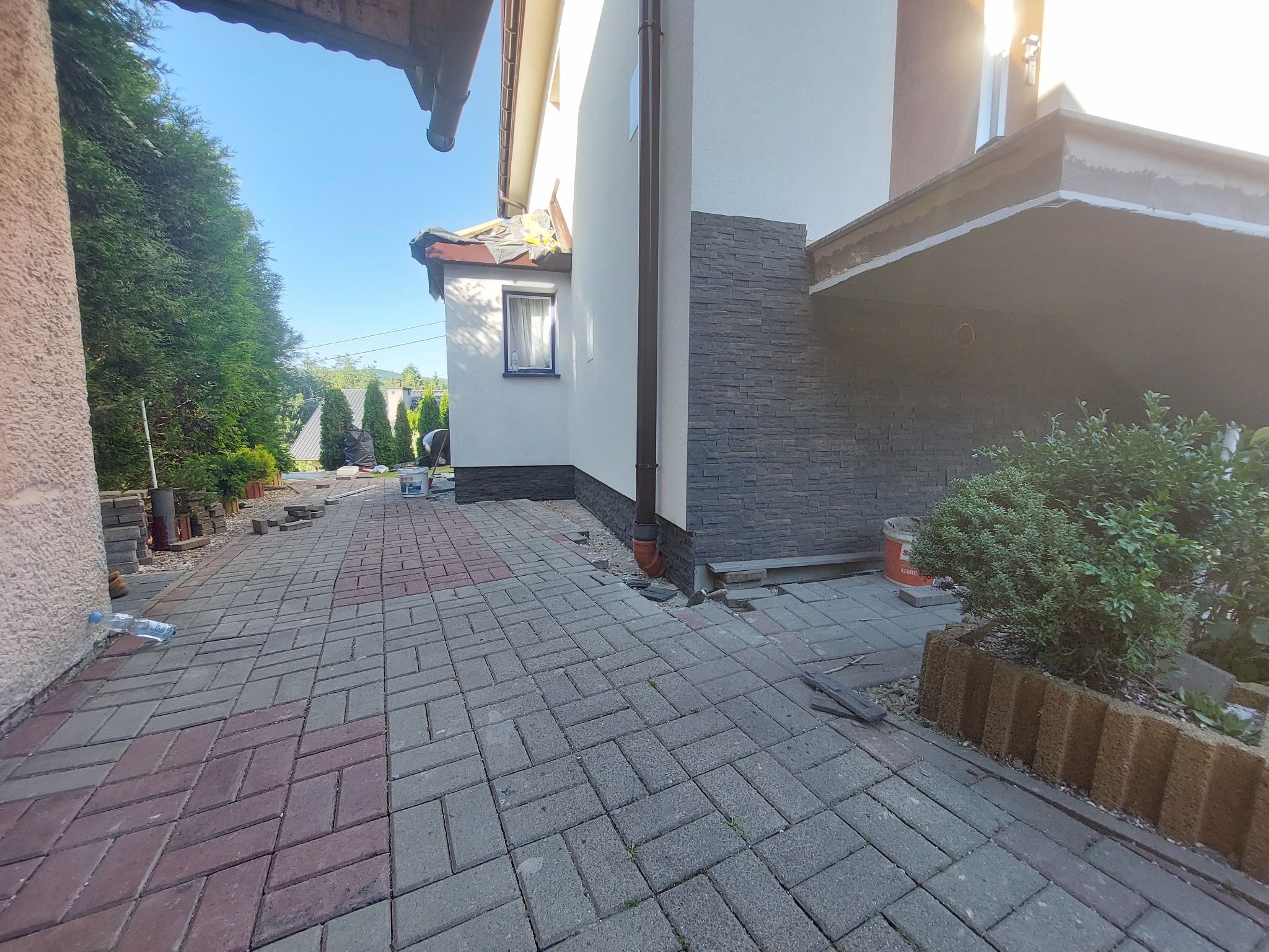 Fragment elewacji domu z dekoracyjną okładziną kamienną, widoczny fragment chodnika z kostki brukowej, prace w toku.