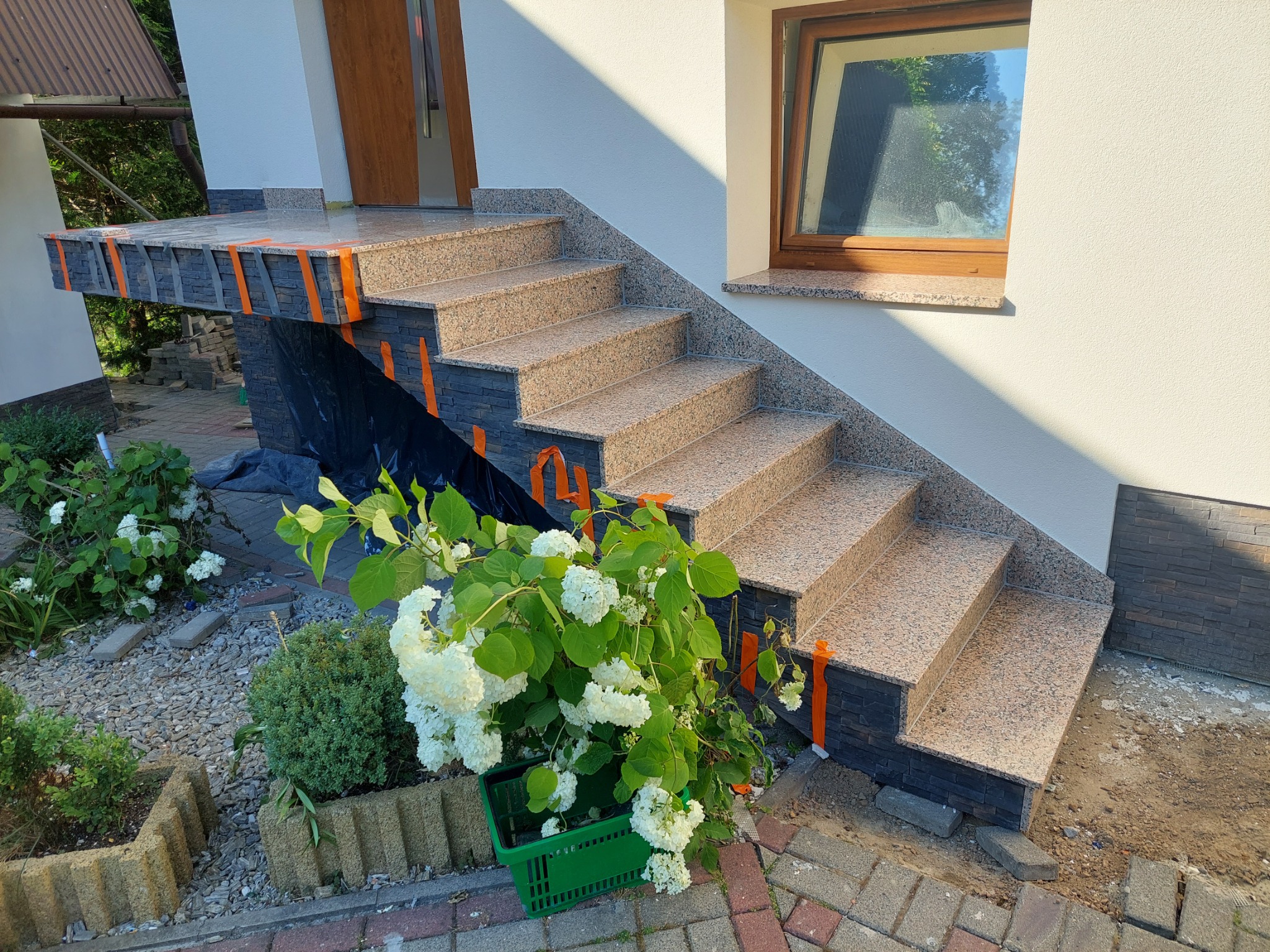 Granitowe schody wejściowe do domu z okładziną z kamienia elewacyjnego, zabezpieczone taśmą malarską, z widoczną roślinnością w donicach.