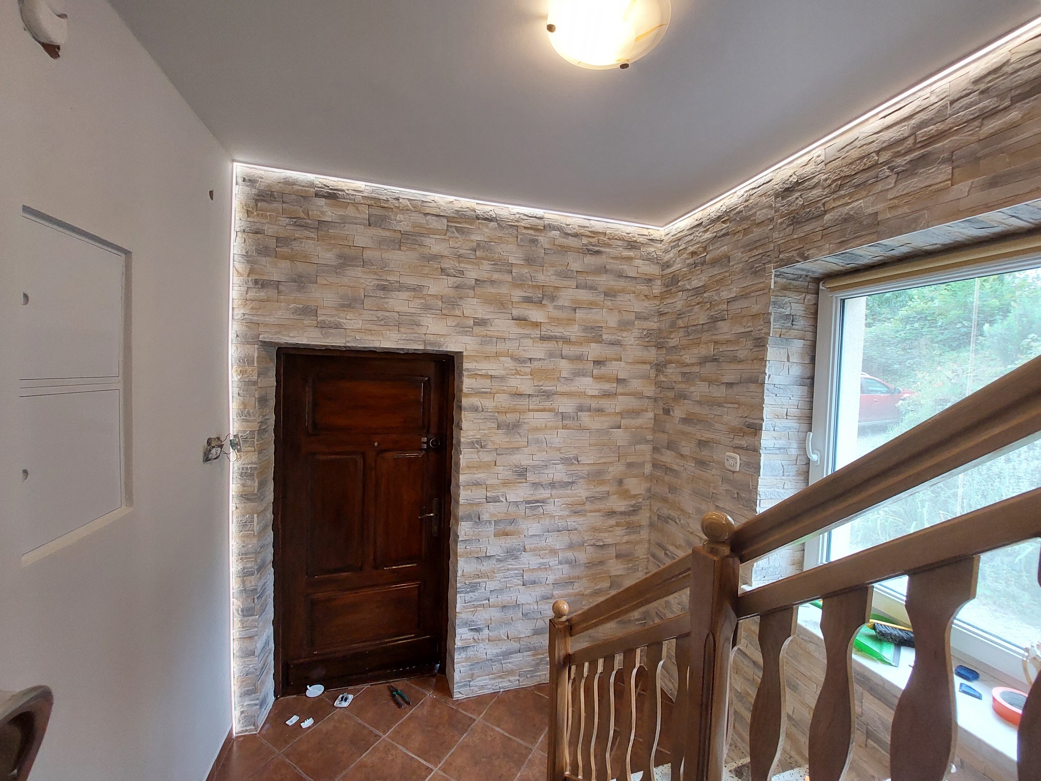 Korytarz po remoncie: ściana wyłożona dekoracyjnym kamieniem, oświetlenie LED pod sufitem, drewniane drzwi i balustrada schodów, brązowa terakota na podłodze, widoczne narzędzia pracy przy drzwiach.