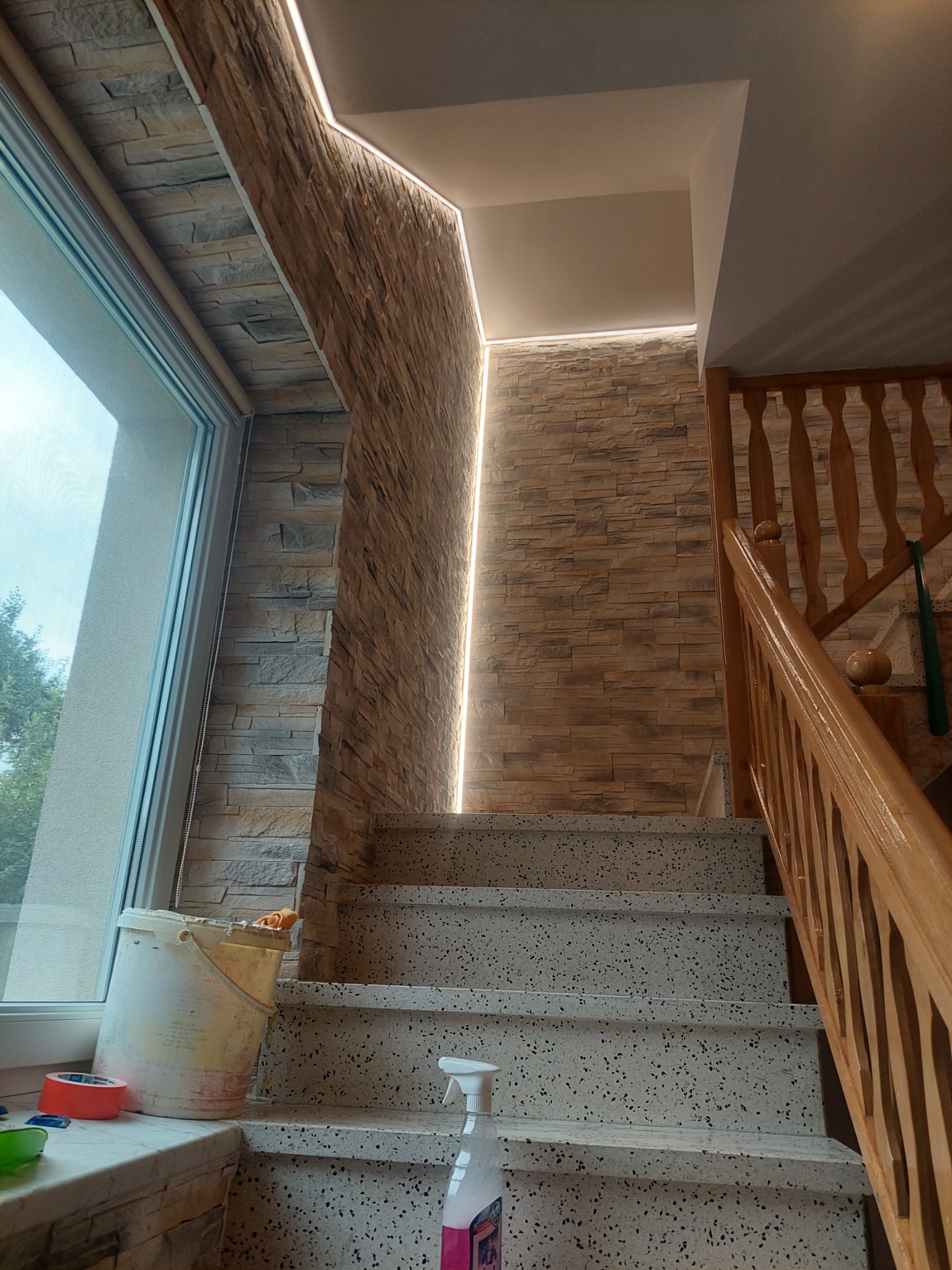 Klatka schodowa z wykończonymi ścianami imitującymi kamień, podświetleniem LED i terazzo na schodach, widoczne narzędzia malarskie i wiadro na parapecie.