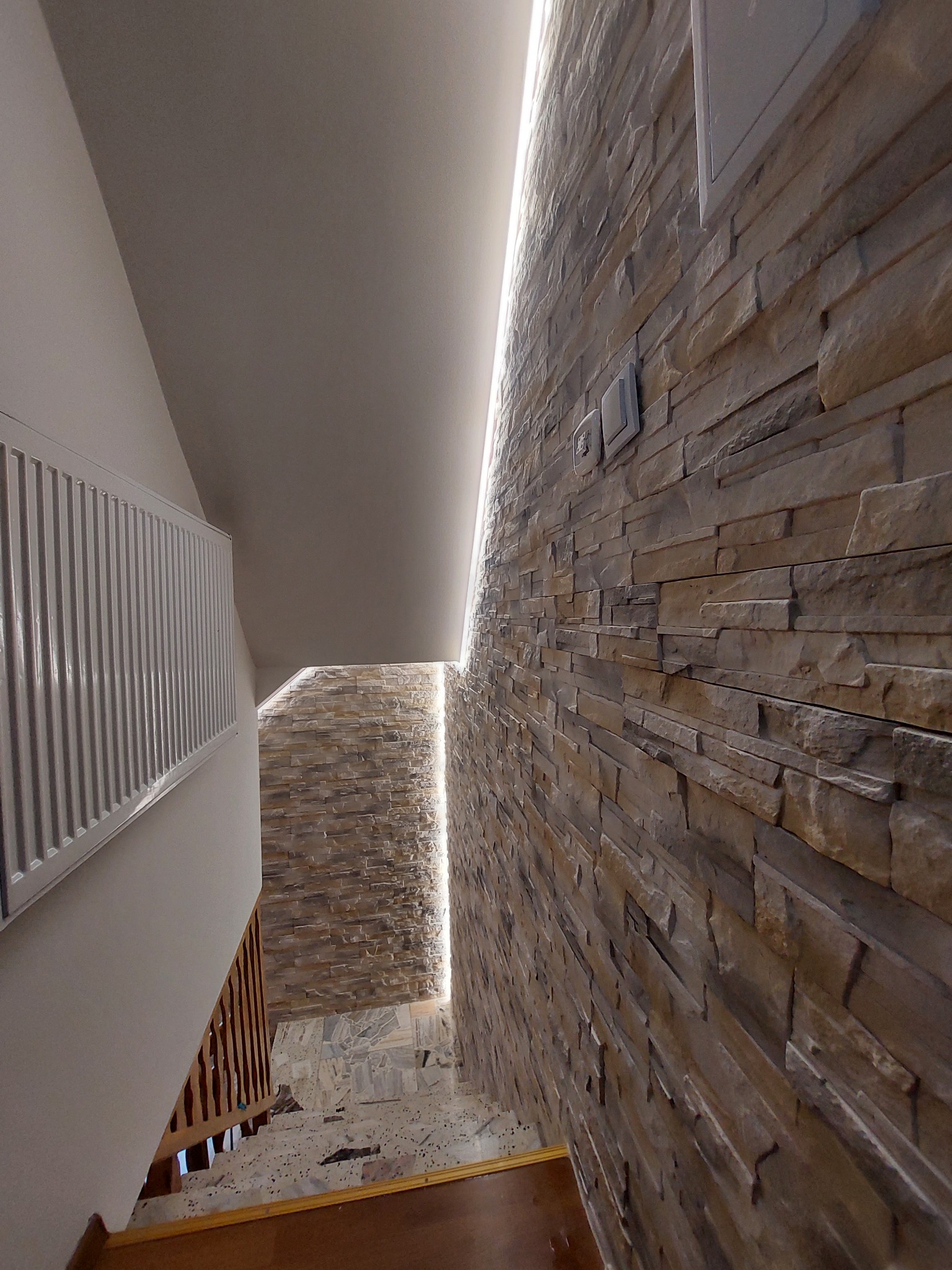 Korytarz ze schodami, ściana wyłożona dekoracyjnym kamieniem w odcieniach brązu i beżu, podświetlona pionową listwą LED, z widocznym grzejnikiem i elementami balustrady.