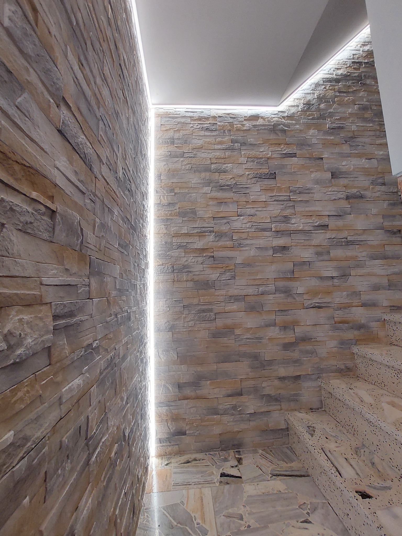 Wąski korytarz wykończony dekoracyjną okładziną kamienną w odcieniach beżu i szarości, oświetlony liniowym światłem LED wzdłuż sufitu i ściany, marmurowe schody z terazzo.
