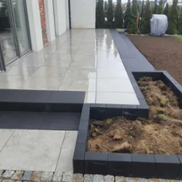 Nowoczesny taras z jasnych, błyszczących płyt, czarne schody i obrzeża, przygotowana rabata kwiatowa z ziemią, w tle tuje i sprzęt ogrodowy pod pokrowcem.