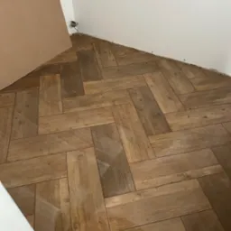 Ułożona podłoga z paneli imitujących drewno w jodełkę, widoczne niedokończone prace przy ścianie i wystające kable elektryczne.