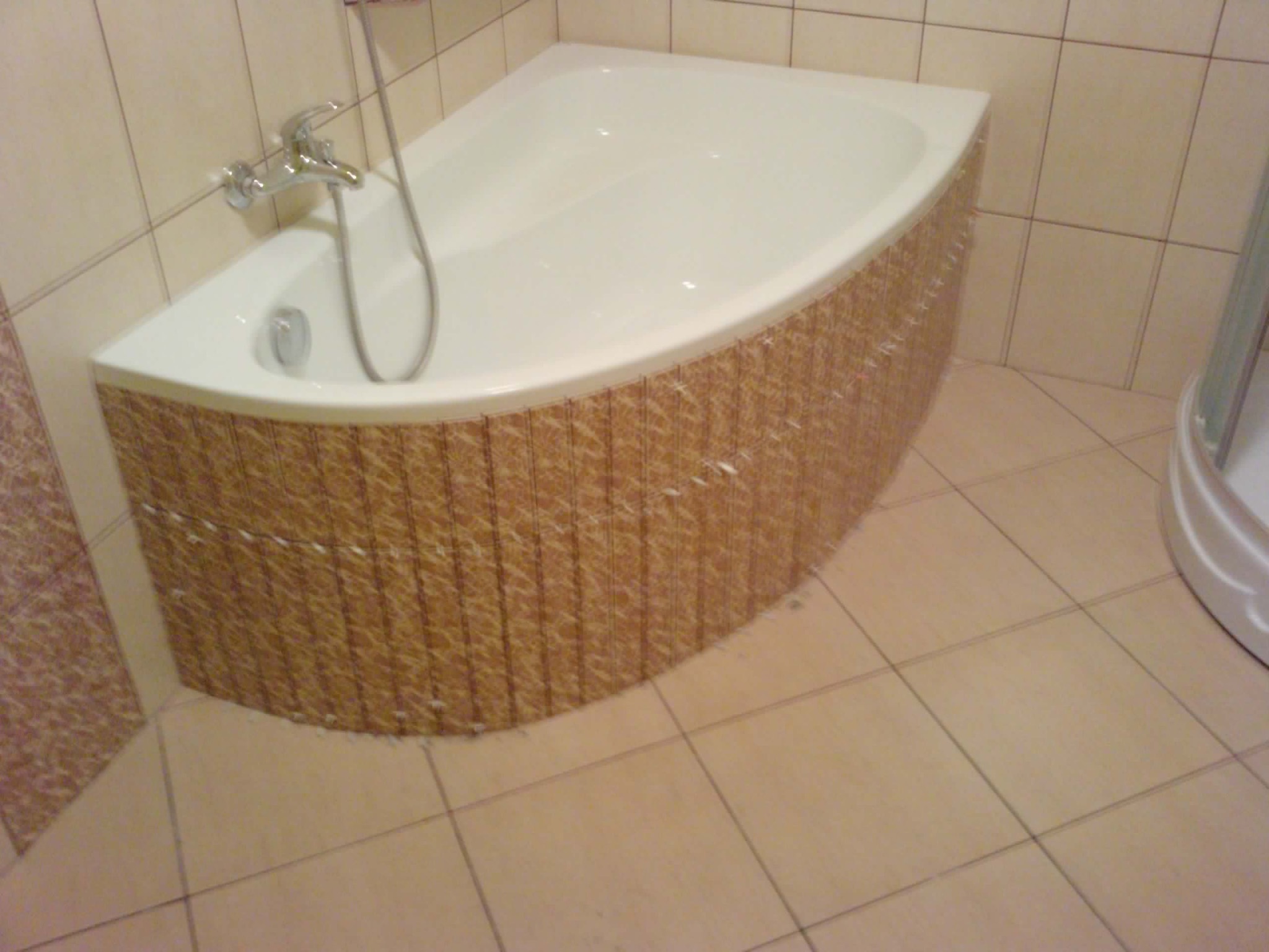 Narożna wanna z obudową z pionowych płytek w odcieniach brązu, chromowana bateria i słuchawka prysznicowa, beżowe płytki podłogowe i ścienne, fragment kabiny prysznicowej.