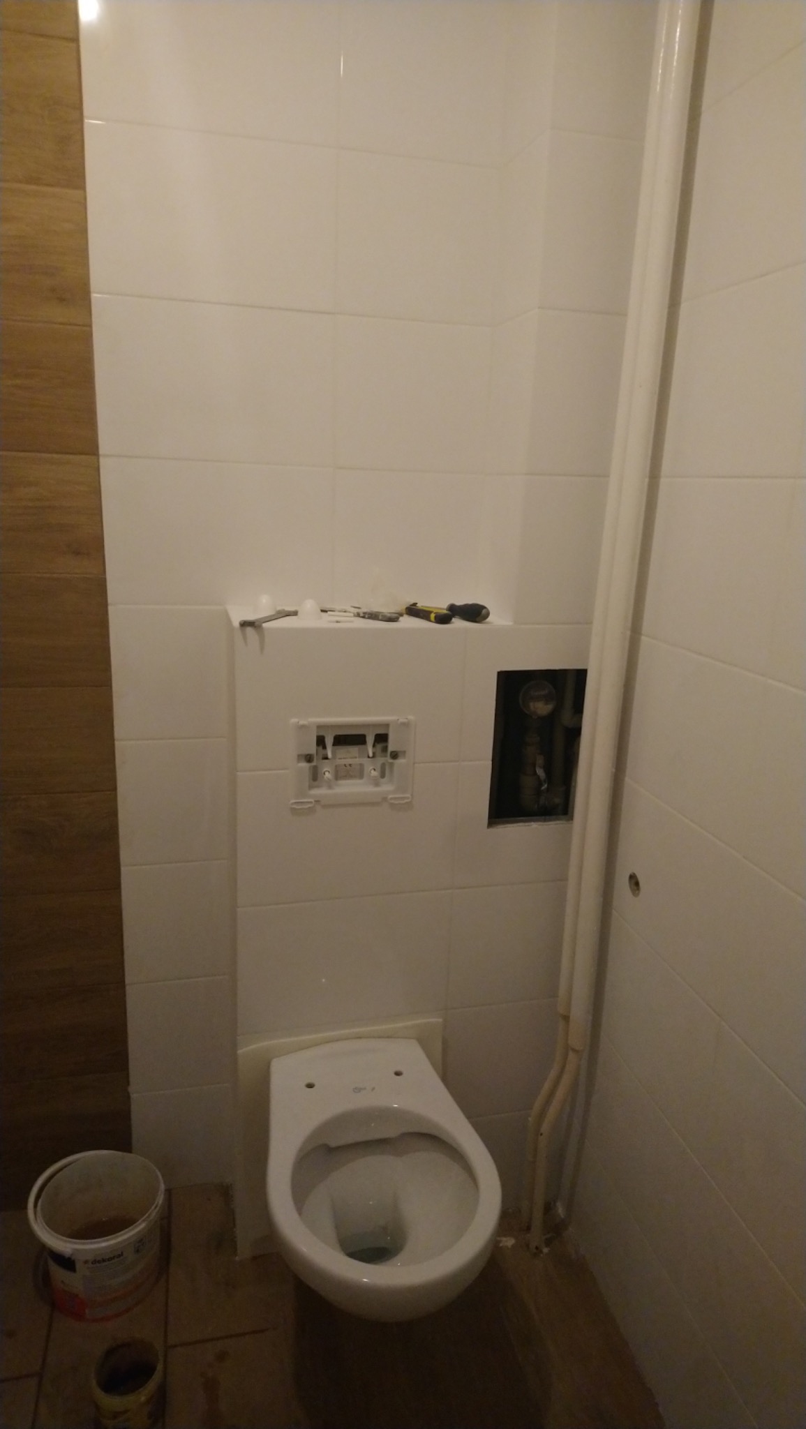 Niezamontowana toaleta podwieszana w trakcie prac wykończeniowych łazienki, widoczne narzędzia na obudowie stelaża, odkryte rury i białe płytki na ścianach.