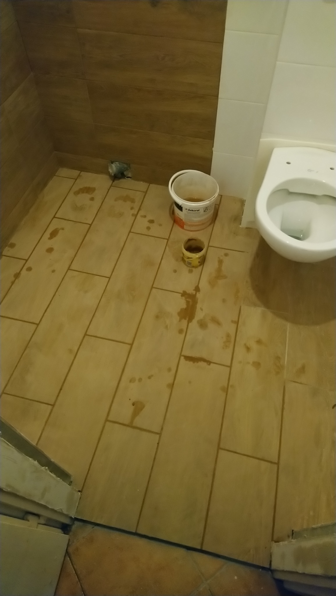Niedbale wykafelkowana podłoga w łazience z plamami po farbie, widoczne wiaderko z farbą Dekoral i puszka po farbie, w tle toaleta i ściany wyłożone brązowymi i białymi płytkami.