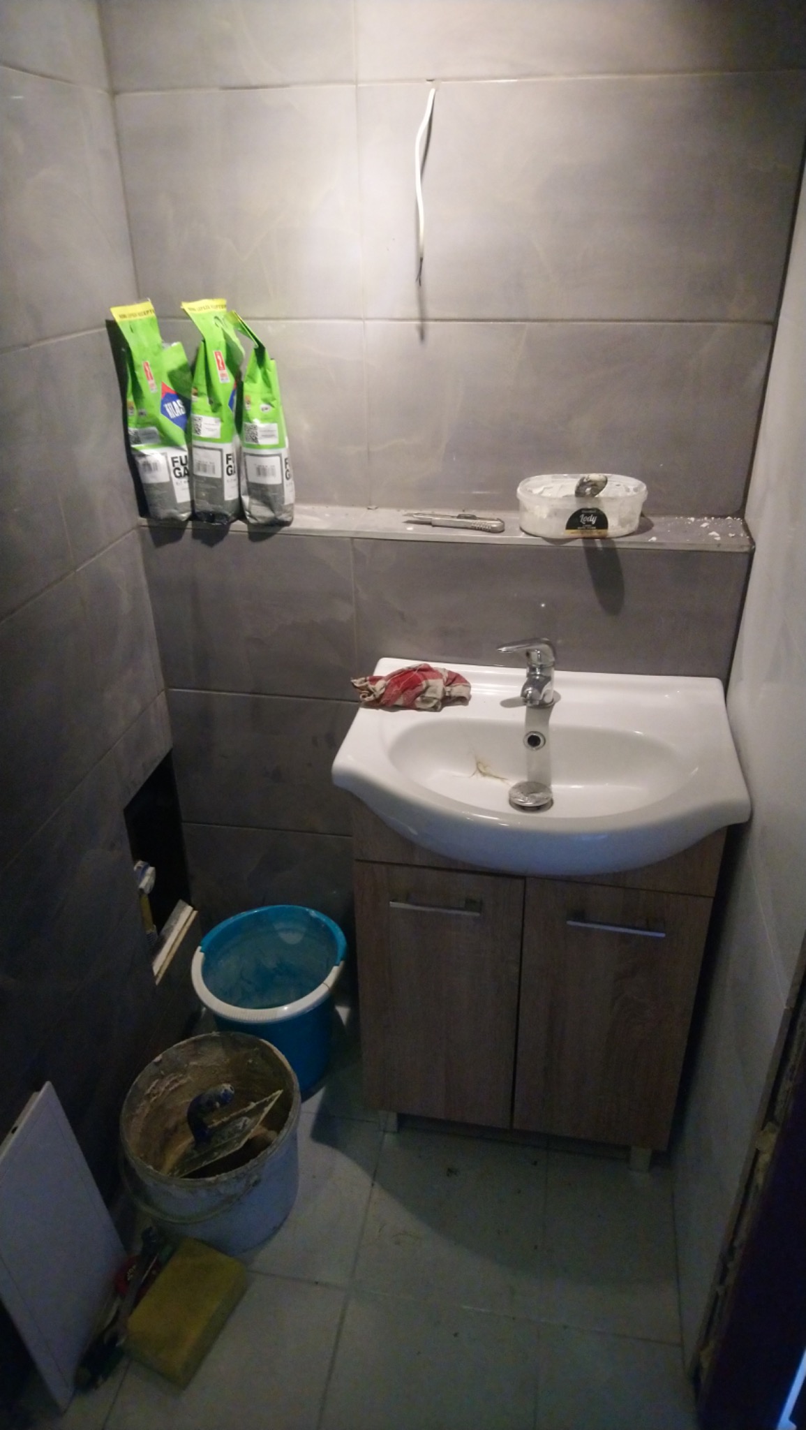 Łazienka w trakcie remontu: szare płytki, umywalka z szafką, worki z fugą, wiadro z zaprawą i narzędzia na podłodze.