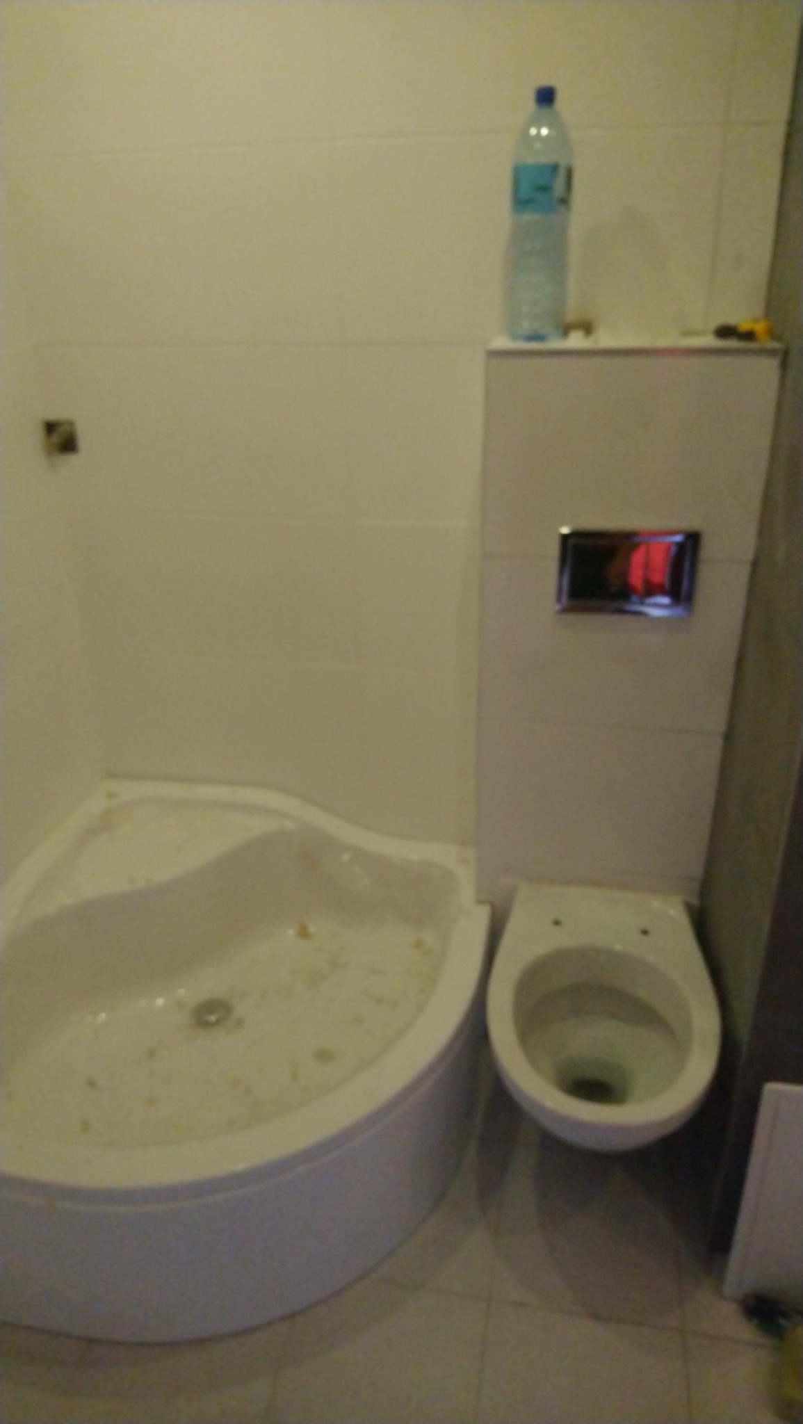 Niedokończona łazienka: narożna kabina prysznicowa z wodą i resztkami po remoncie, wisząca toaleta, płytki na ścianach, butelka wody i narzędzie na zabudowie geberitu.