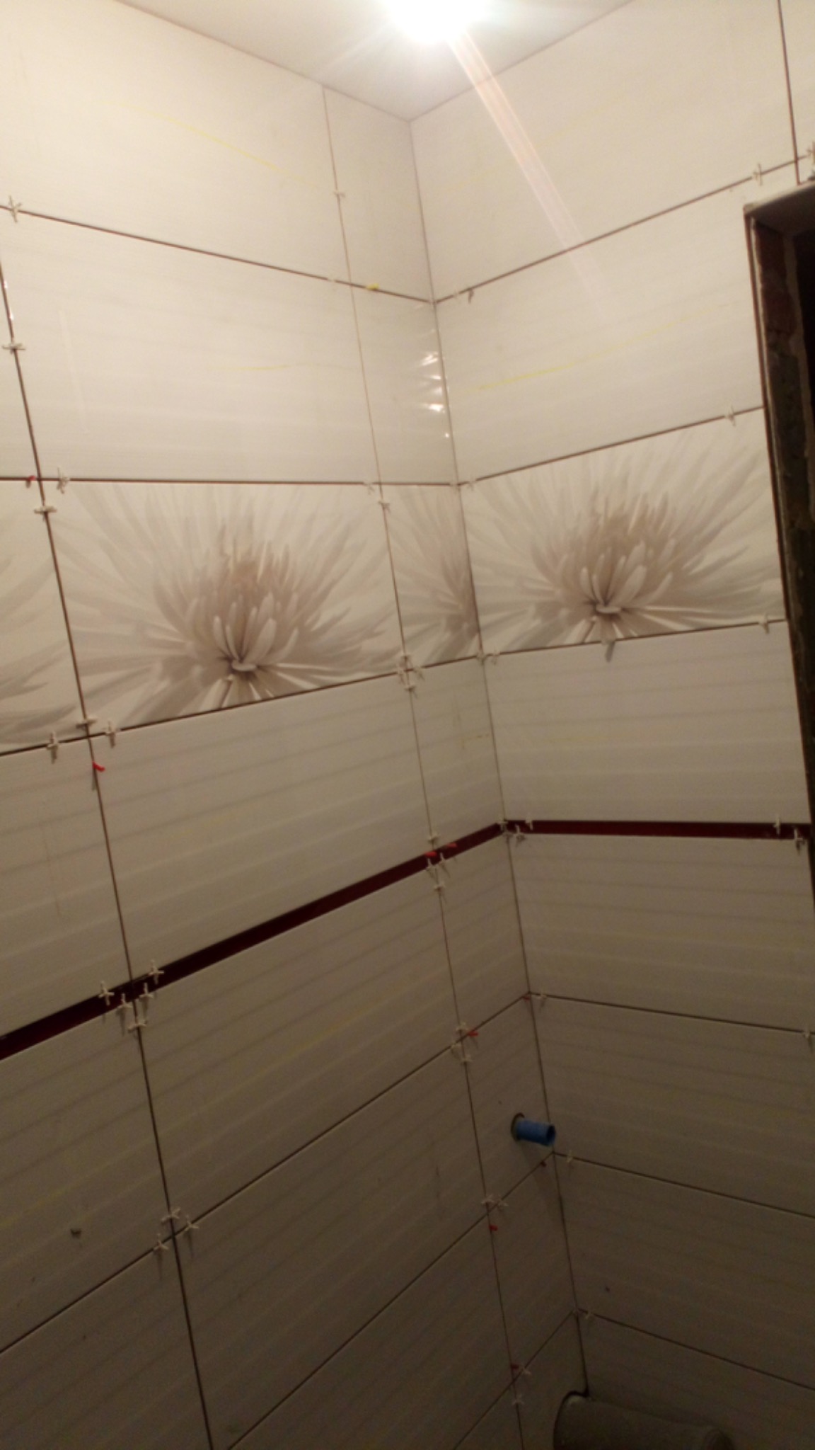 Ściany łazienki wyłożone białymi płytkami z poziomym motywem kwiatowym, widoczne krzyżyki dystansowe i fragment niebieskiej rurki.