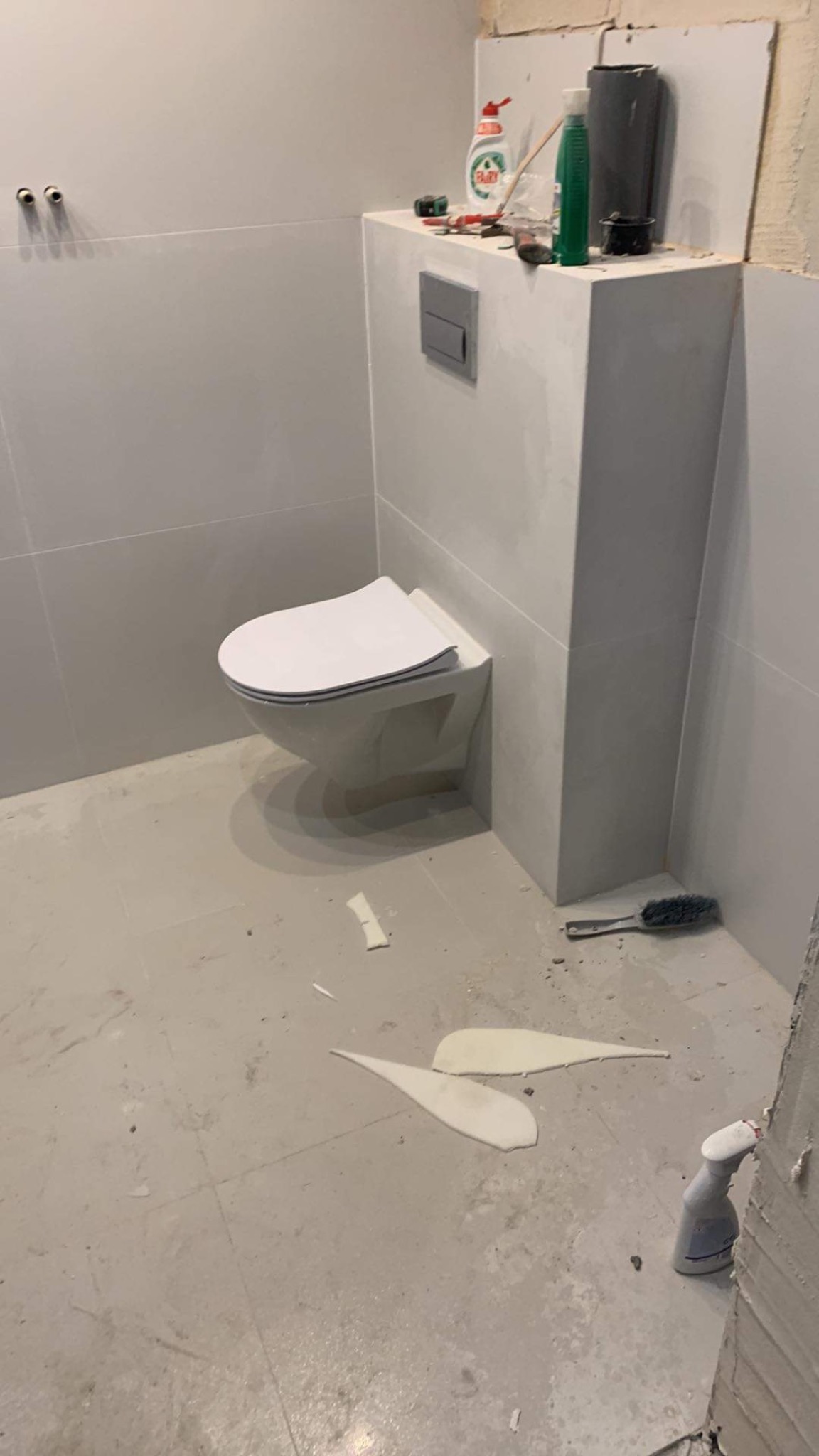 Świeżo zamontowana toaleta podwieszana w surowej, szarej łazience w trakcie prac wykończeniowych, widoczne narzędzia i resztki materiałów budowlanych na podłodze.