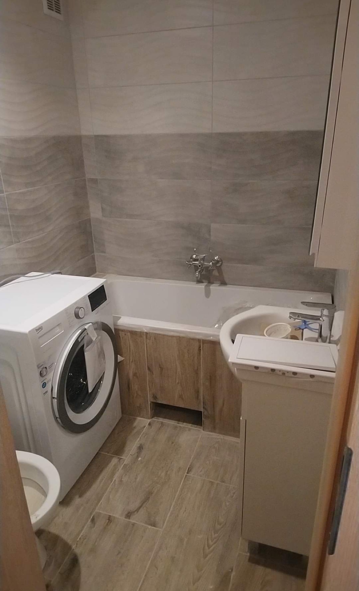 Ukończona łazienka z płytkami imitującymi drewno na podłodze i obudowie wanny, pralka Beko obok toalety, umywalka z szafką w rogu, szare płytki na ścianach z subtelnym wzorem fal.
