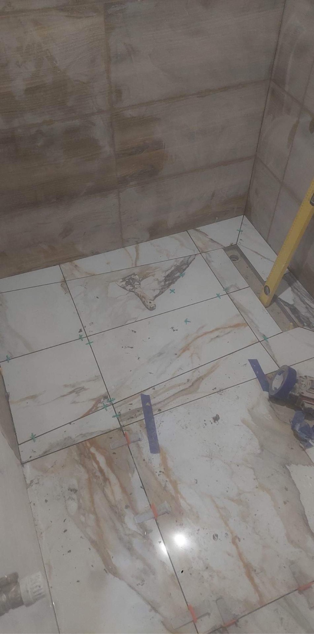 Fragment łazienki w trakcie prac wykończeniowych: białe płytki podłogowe z marmurkowym wzorem, poziomica przy ścianie, widoczne elementy montażowe i zabrudzenia.