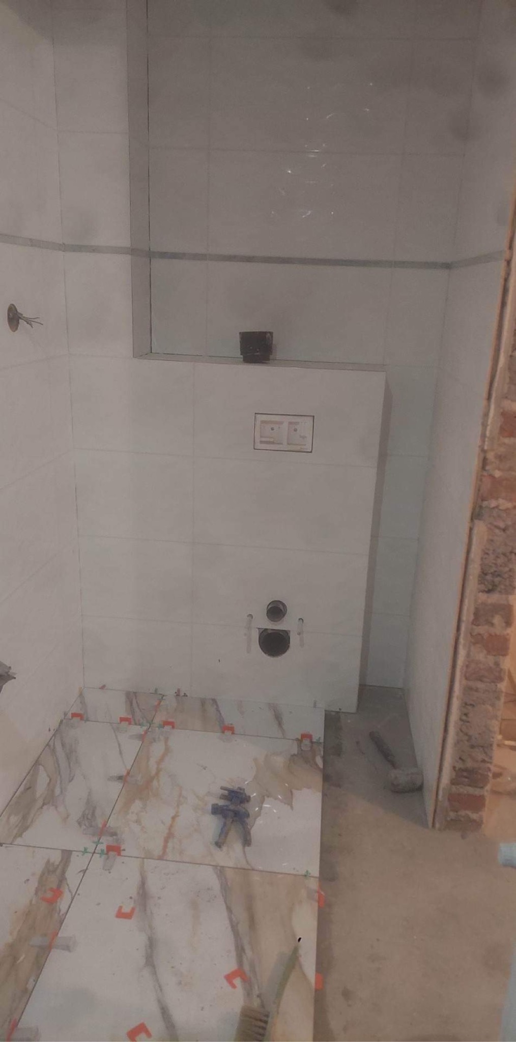 Białe płytki ścienne i podłogowe z marmurkowym wzorem w trakcie montażu w łazience, widoczne plastikowe krzyżyki dystansowe i narzędzie do płytek.