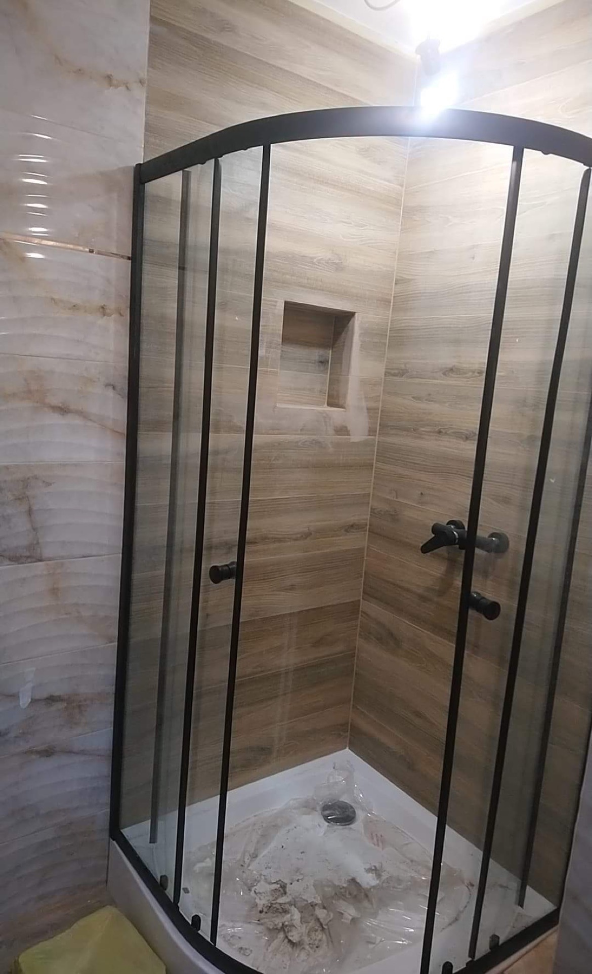 Nowoczesna kabina prysznicowa z czarnymi profilami, zamontowana w narożniku łazienki wyłożonej płytkami imitującymi drewno, z widoczną wnęką w ścianie i pozostałościami po montażu w brodziku.