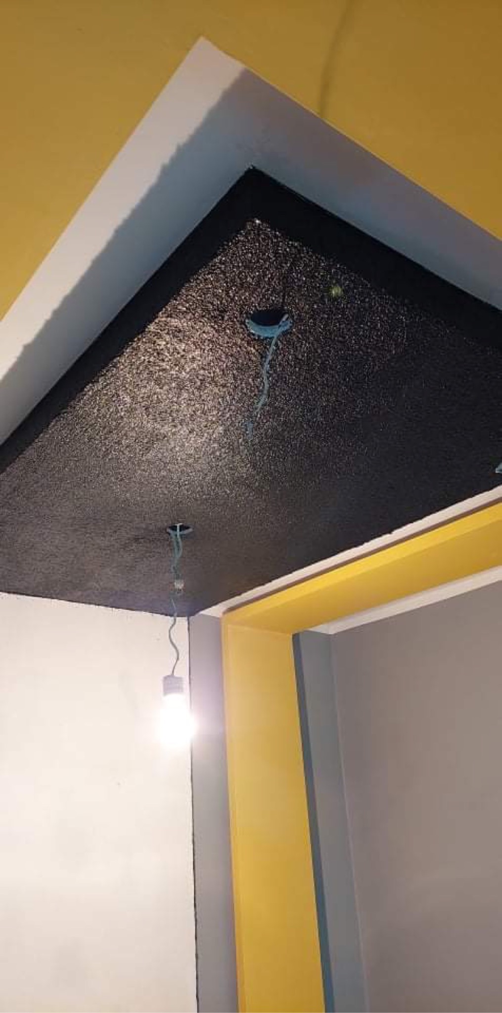 Czarny, teksturowany sufit podwieszany z wystającymi przewodami elektrycznymi i żarówką, otoczony żółtymi i białymi ścianami.